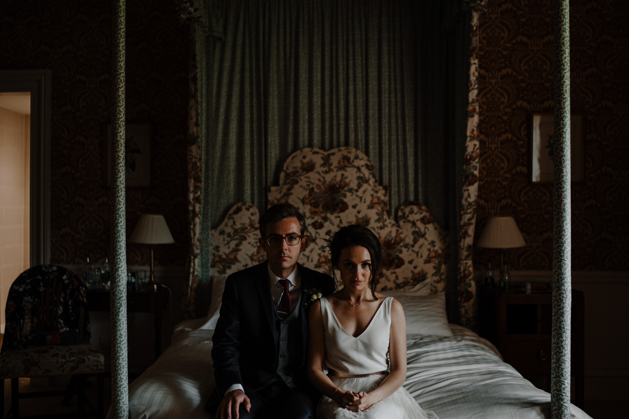 A man and woman in wedding attire sitting on a bed in a vintage-style room with floral wallpaper and draperies.