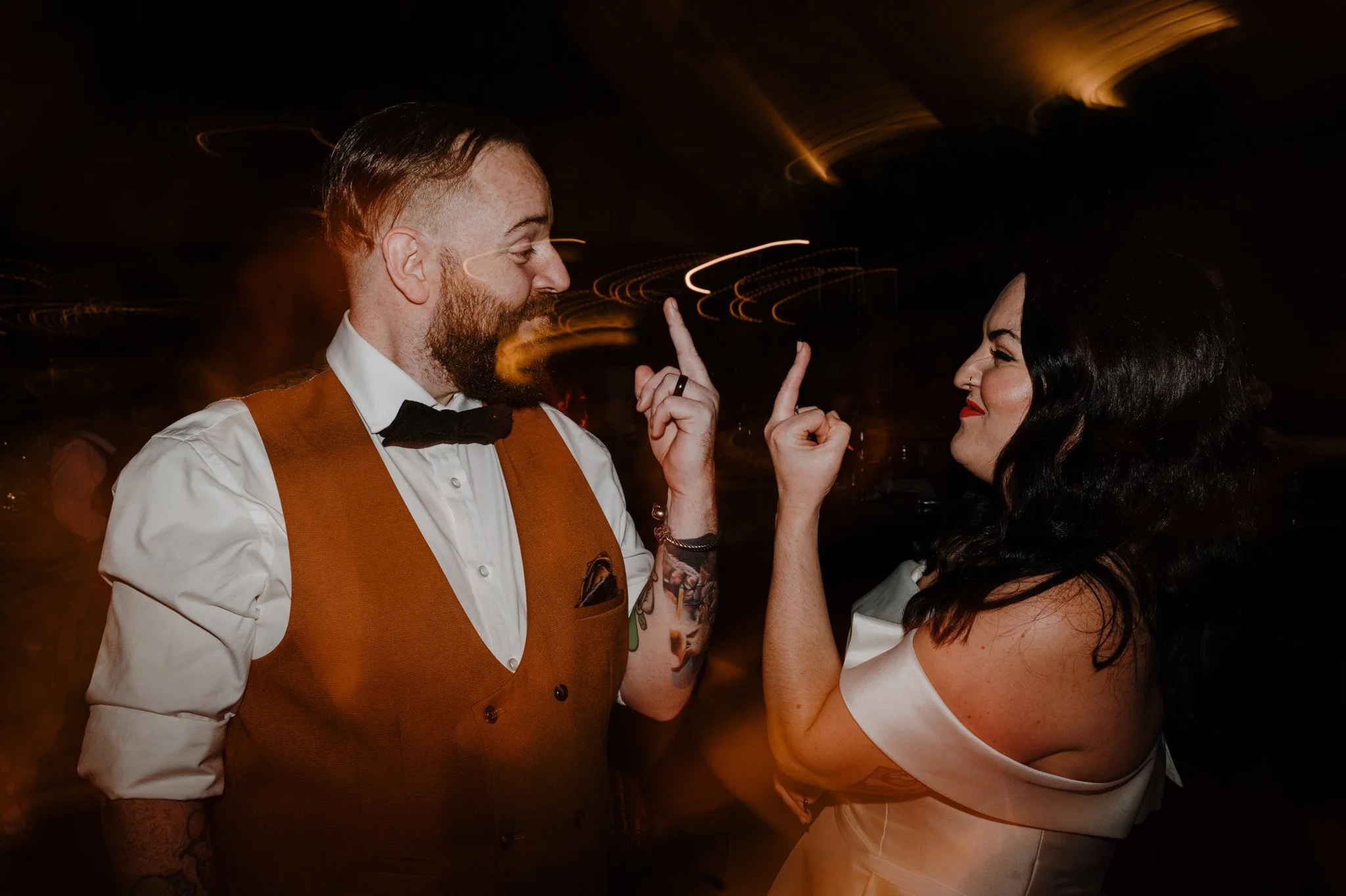 A man with a beard and tattoos, dressed in a white shirt, black bow tie, and brown vest, and a woman with dark hair and red lipstick, are smiling and pointing their index fingers at each other in a dark, lively setting with warm lighting.