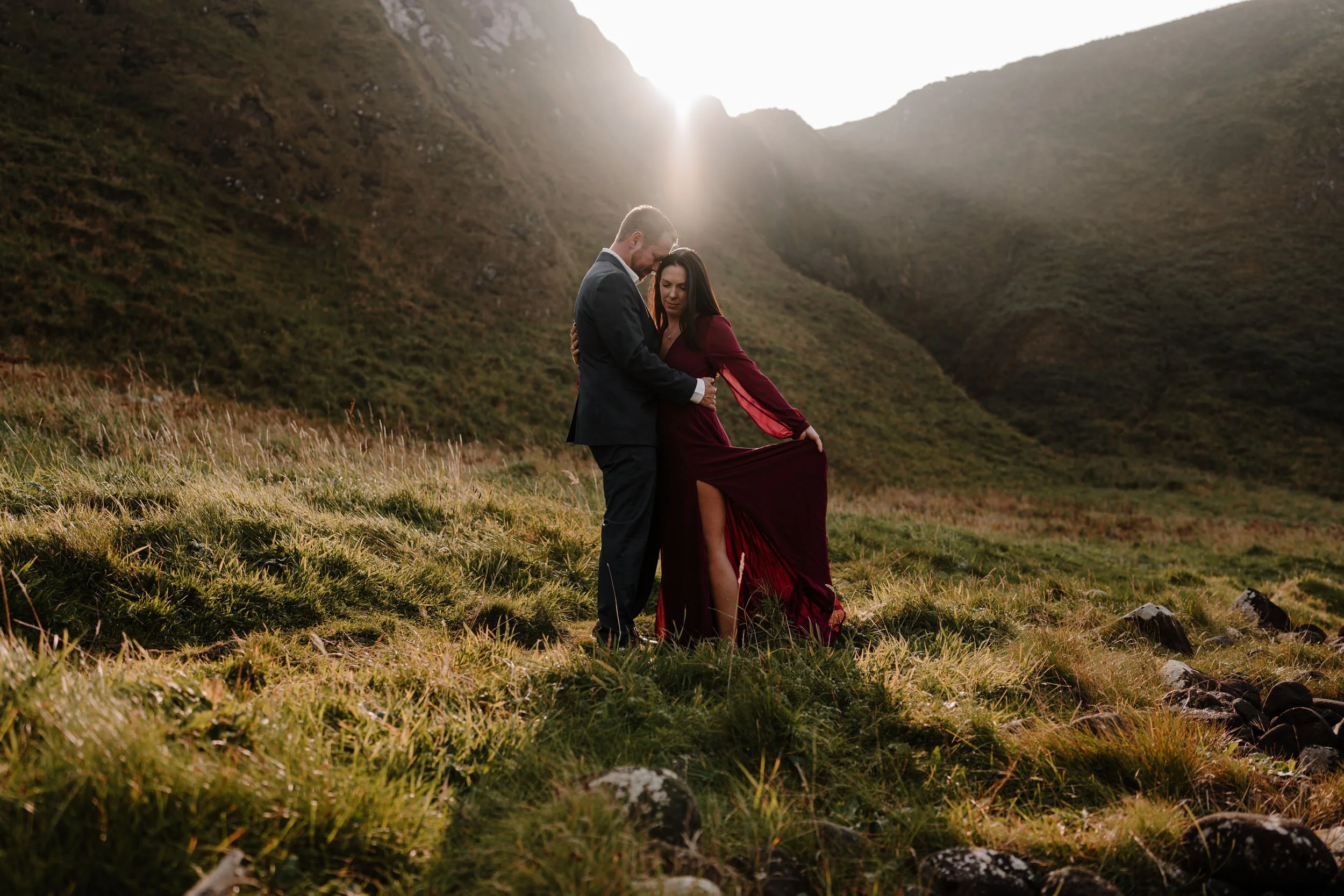 couple embrace in sunlight at kinbane castle for their couples adventure session in spring. woman wears a maroon dress