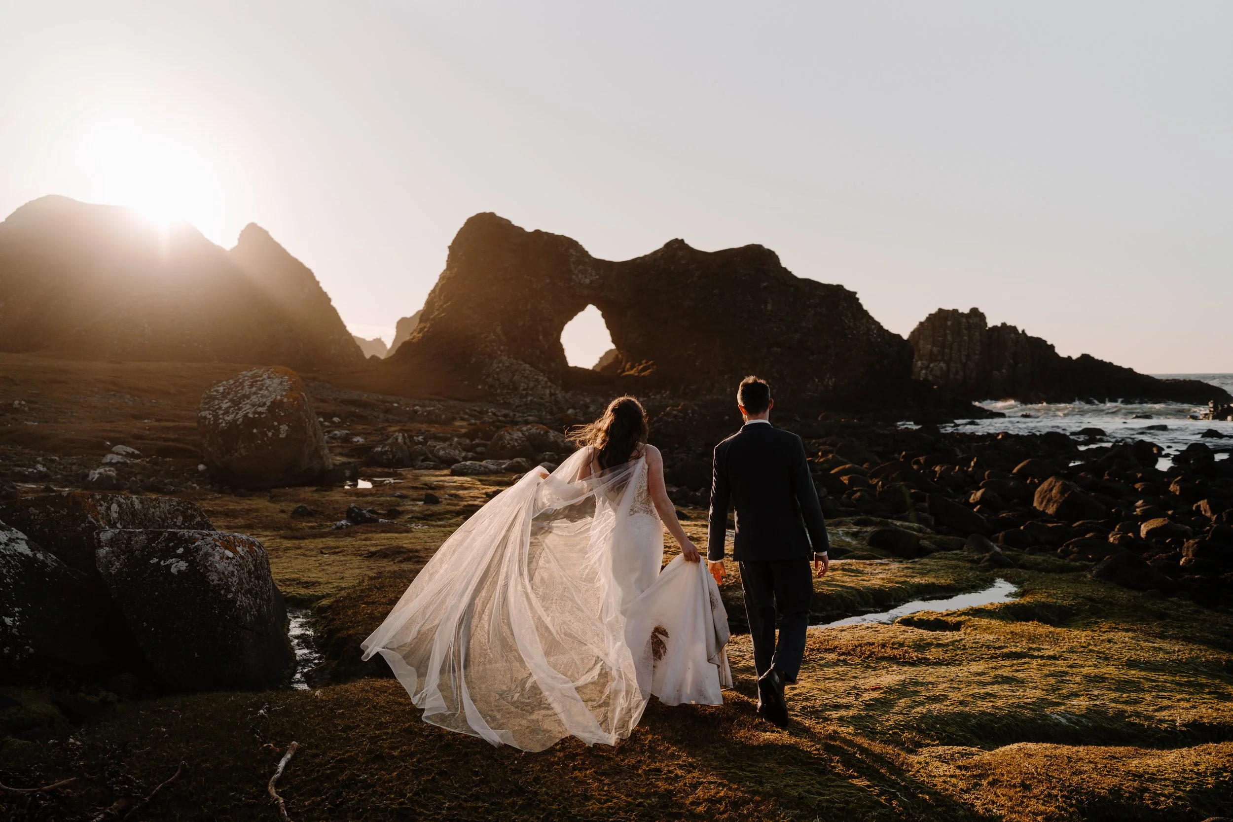 A bride and groom walking on rocky terrain with short grass, holding hands, with a natural rock arch formation and the ocean in the distance, during sunset.