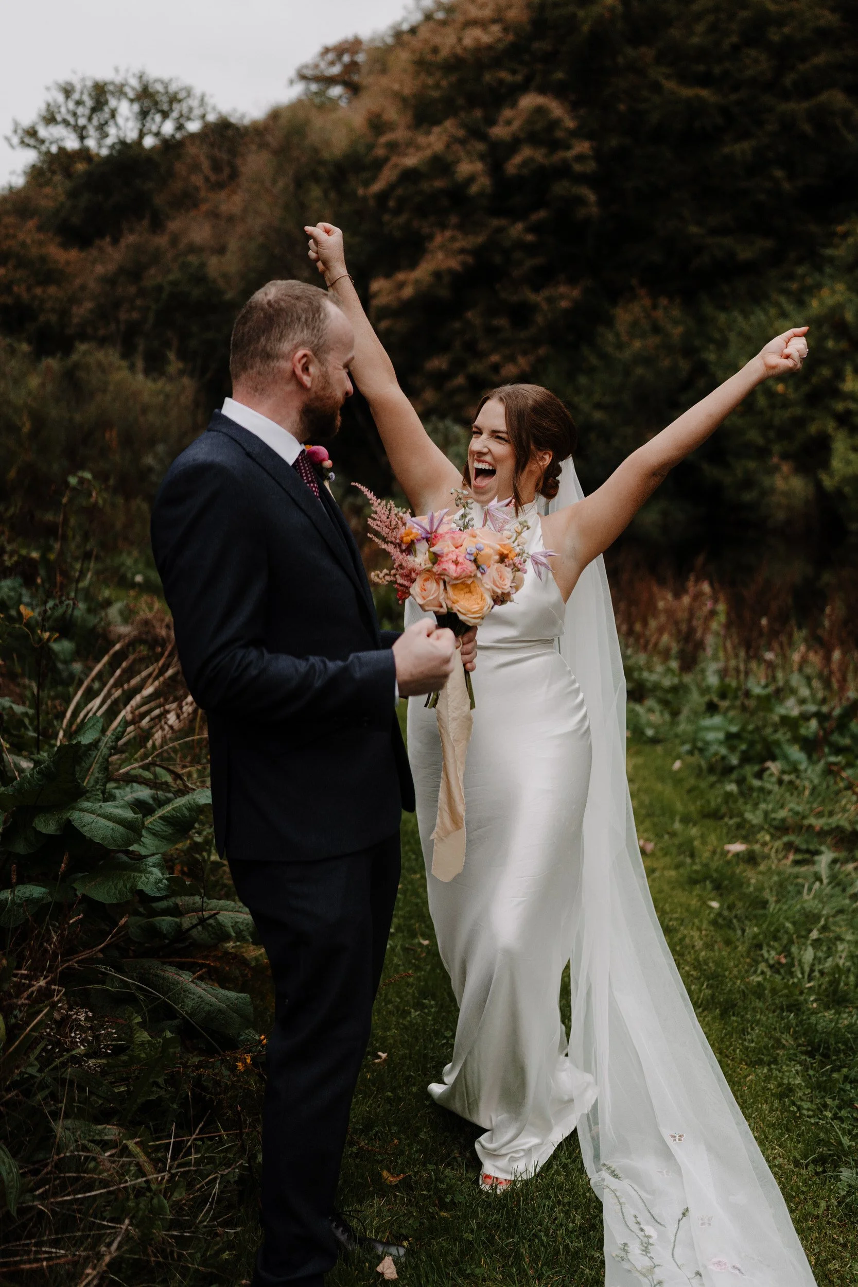 A bride and groom celebrating outdoors during their wedding, with the bride joyfully raising her arms and holding a bouquet, and the groom smiling at her, set against a backdrop of trees with autumn foliage.