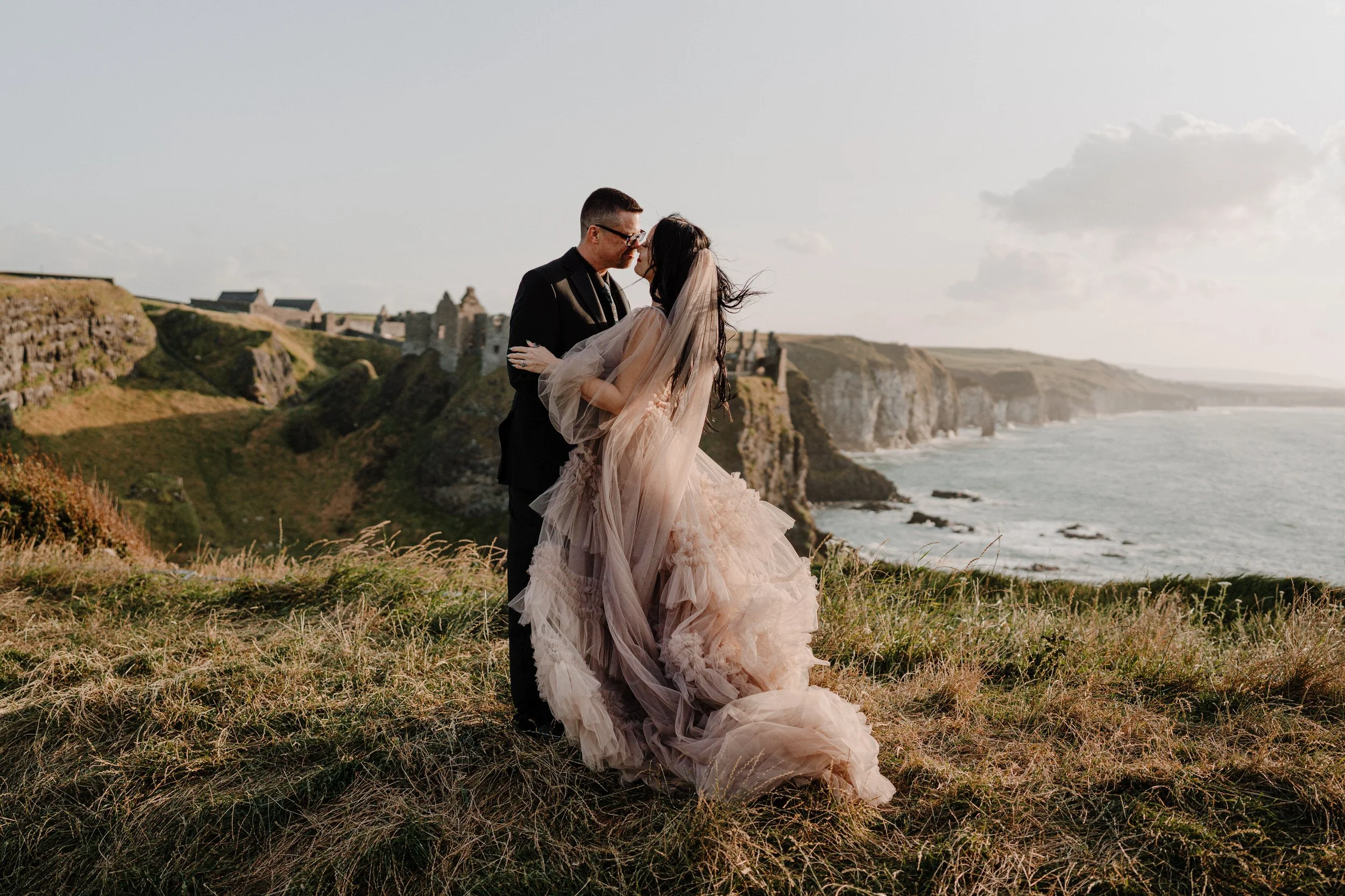 A eloping bride and groom dressed in flowy wedding attire standing on a grassy hill on the cliff edge with Dunluce Castle in the distance. overlooking the ocean and cliffs, embracing and appearing to share a kiss.
