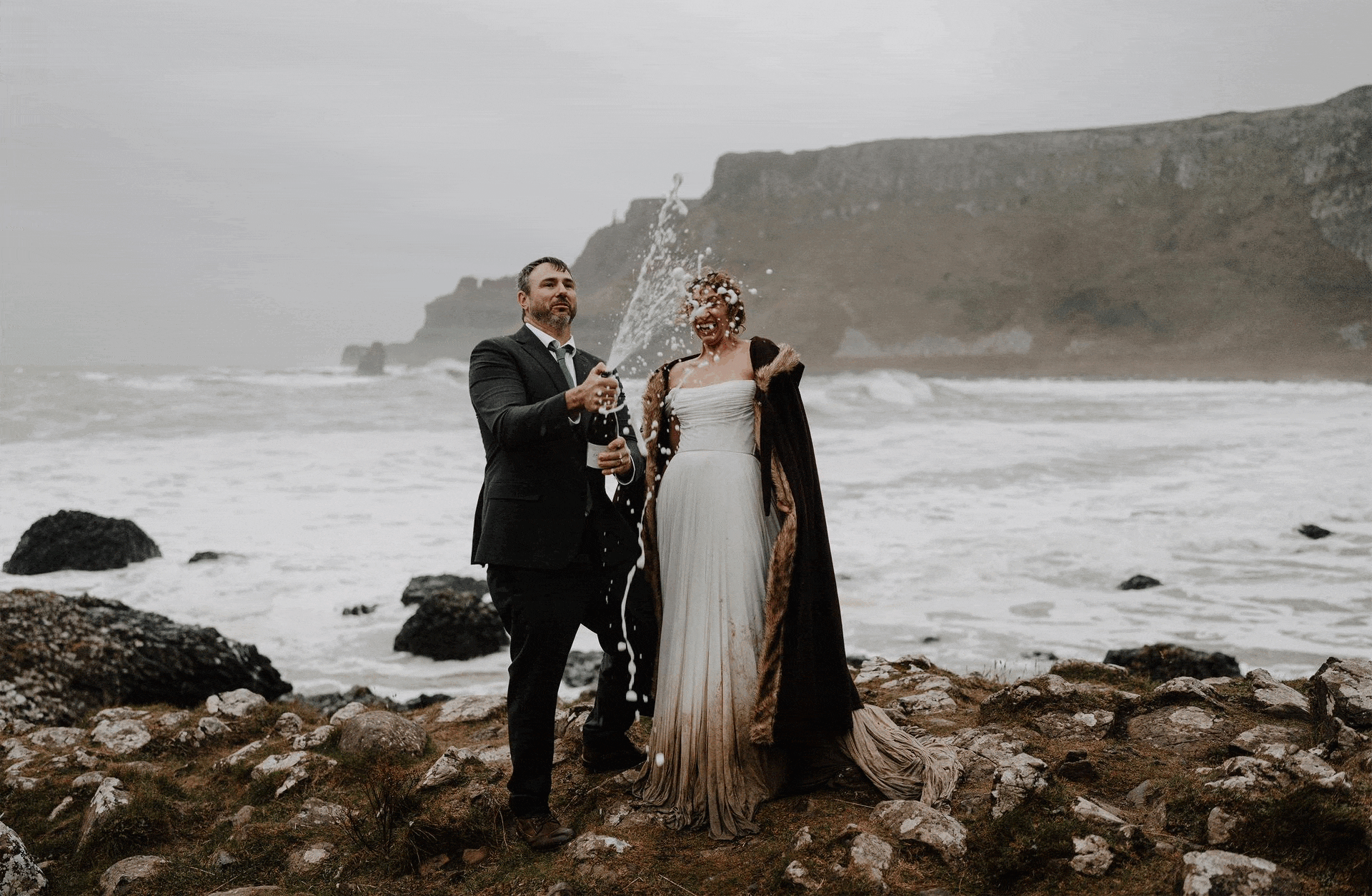 A newlywed couple celebrating on a rocky beach with cliffs in the background. The groom wearing a black suit and tie is holding a champagne bottle, spraying champagne on the bride, who is smiling and wearing a flowing gown with a cape. The scene is overcast with ocean waves crashing behind them.