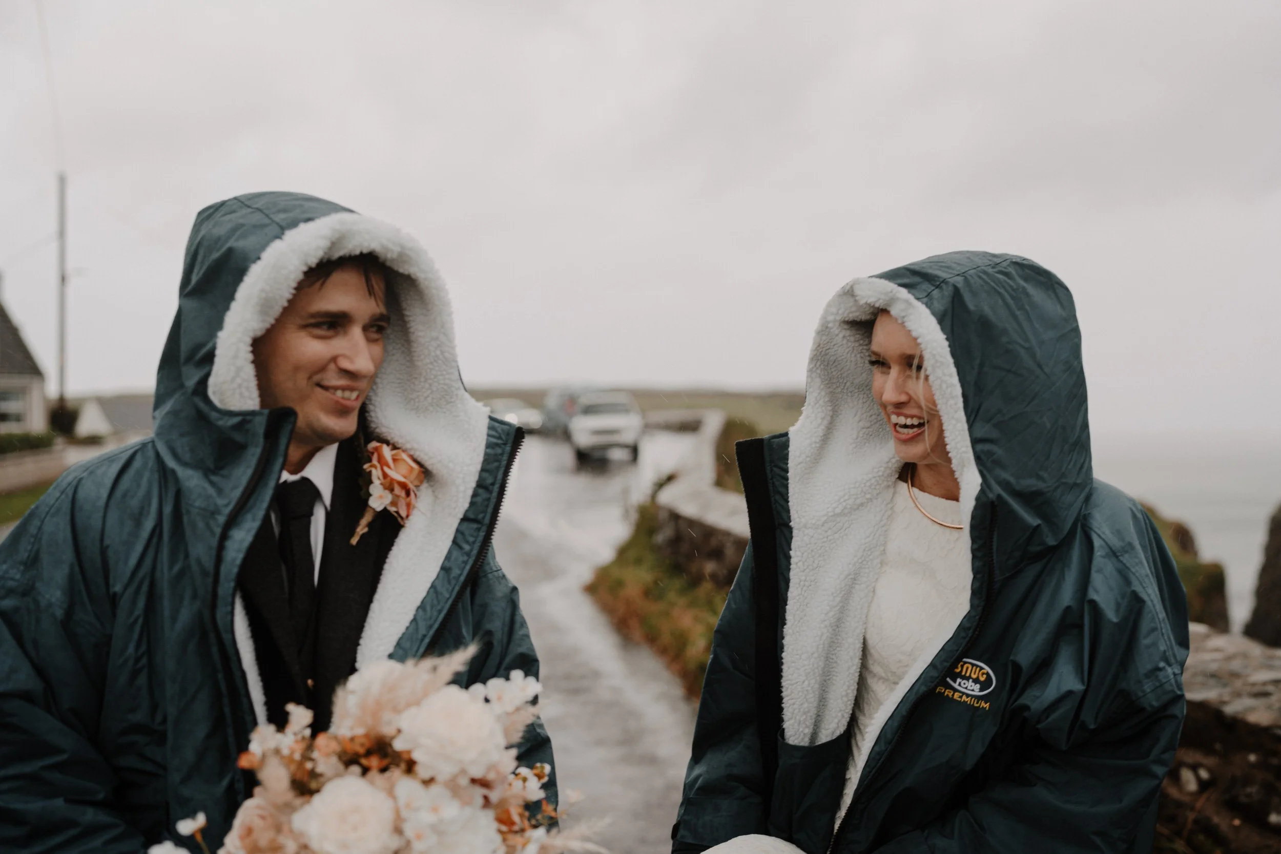 A smiling man and woman wearing hooded jackets with fleece lining outdoors on a cloudy day, with cars and a house in the background.