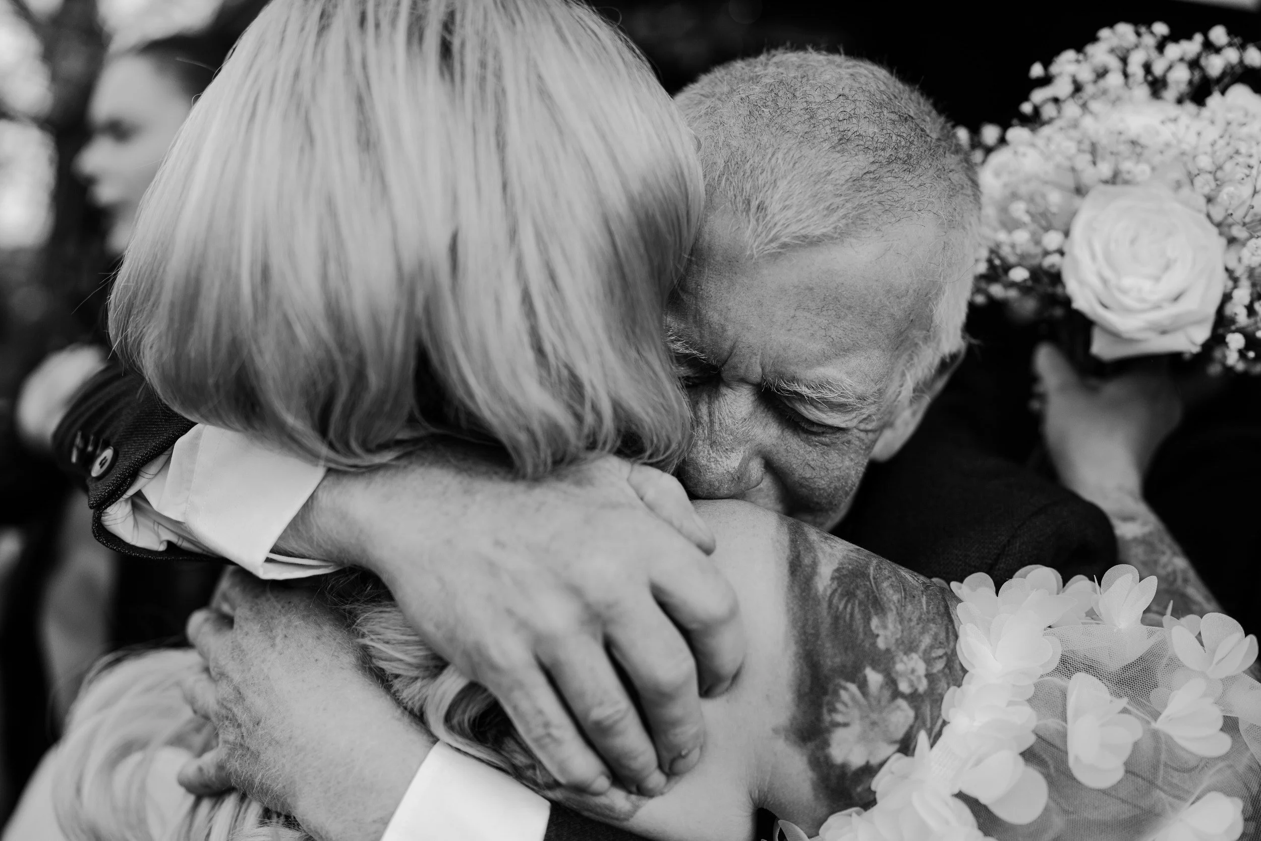 Two people hugging lovingly, with one person having gray hair and the other having a shaved head, surrounded by flowers, in a black and white photo.