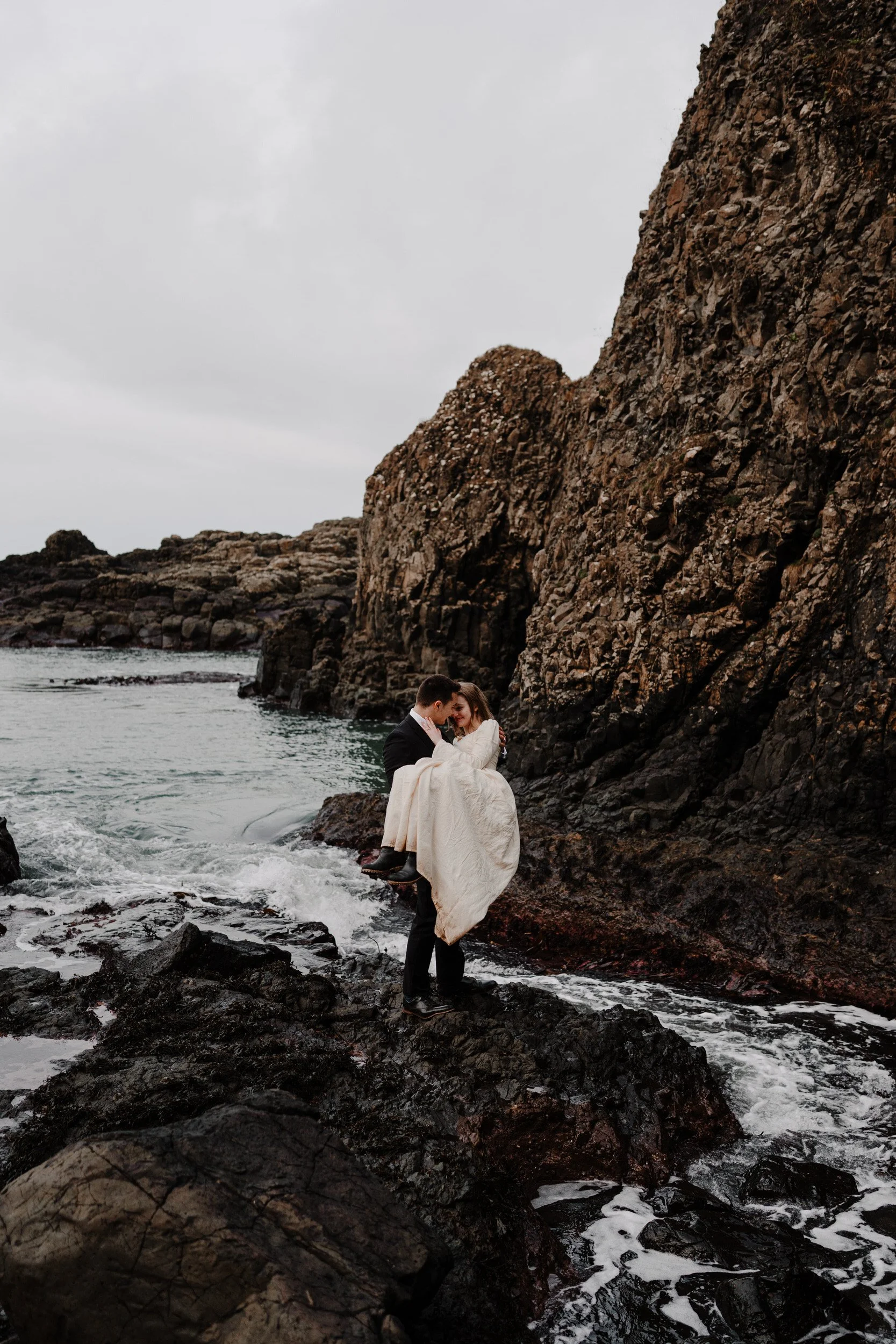 A couple in wedding attire by the ocean on a rocky shoreline, with the groom carrying the bride who is wrapped in a white blanket.