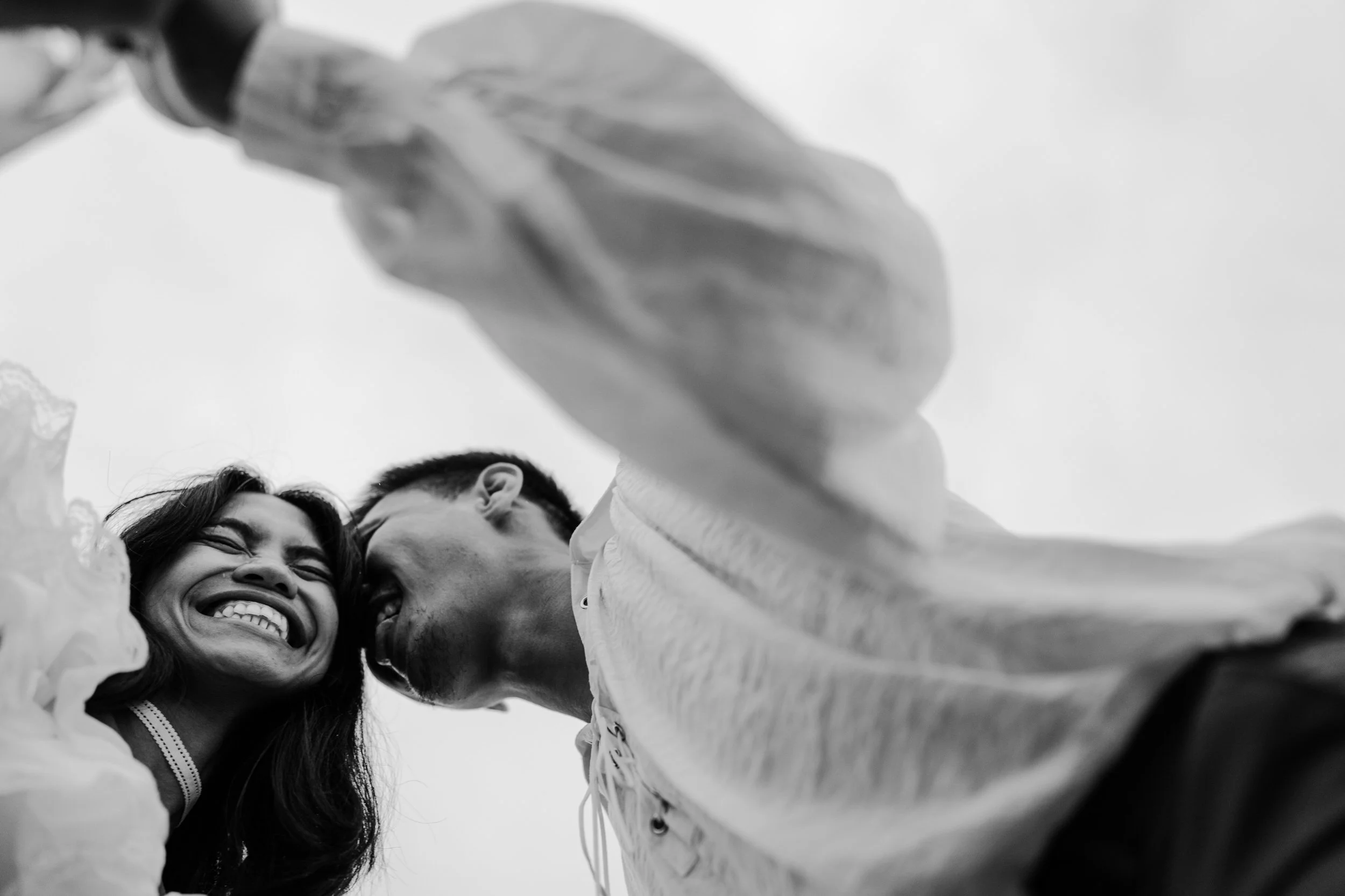 A black and white photo of a woman smiling with her eyes closed, close to a man, both embracing and leaning toward each other, with another person's arm reaching towards the camera in the foreground.
