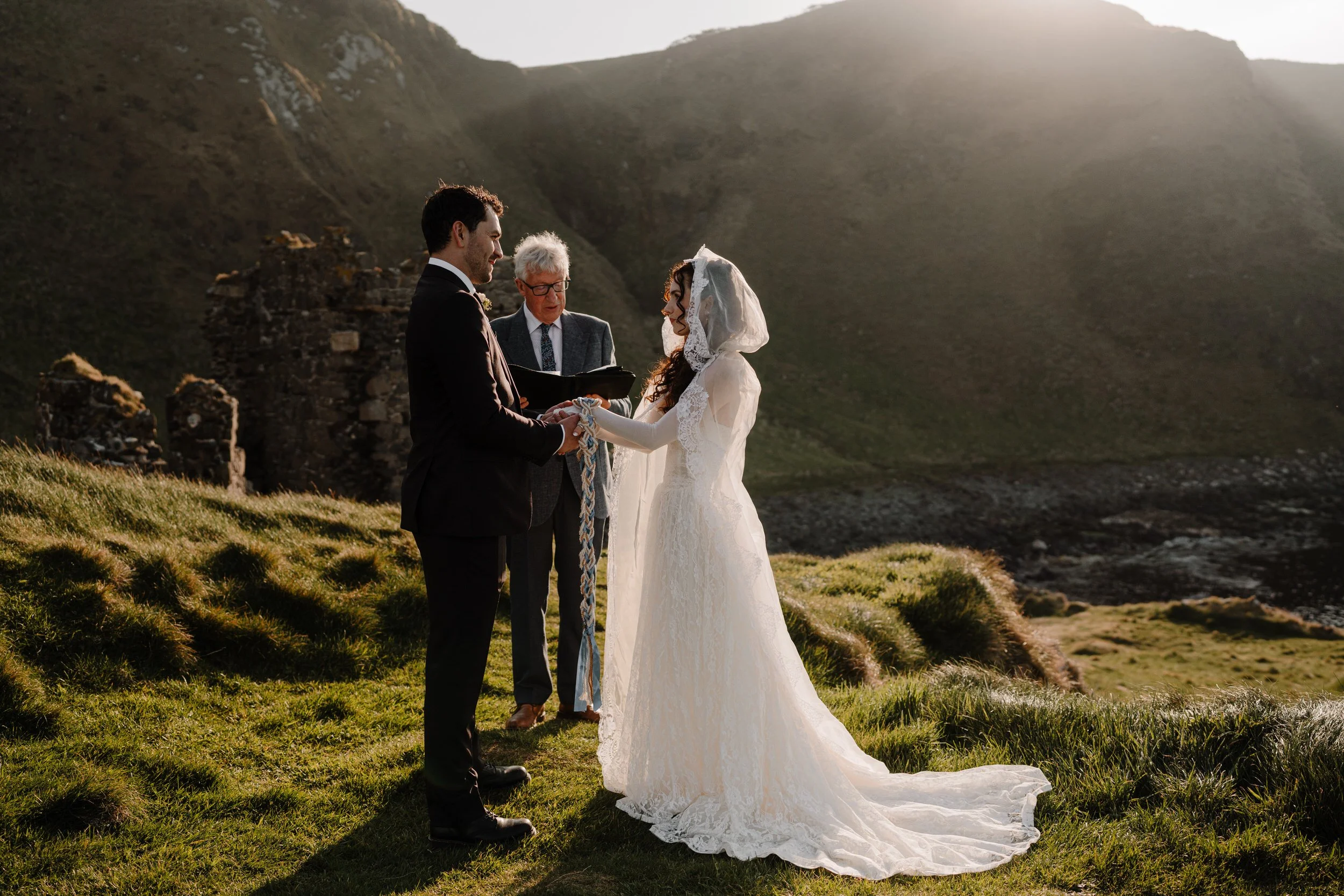 handfasting ceremony for an irish elopement at kinbane castle couple stand in sunlight on the green cliff with their hands bound in a celtic handfasting cord
