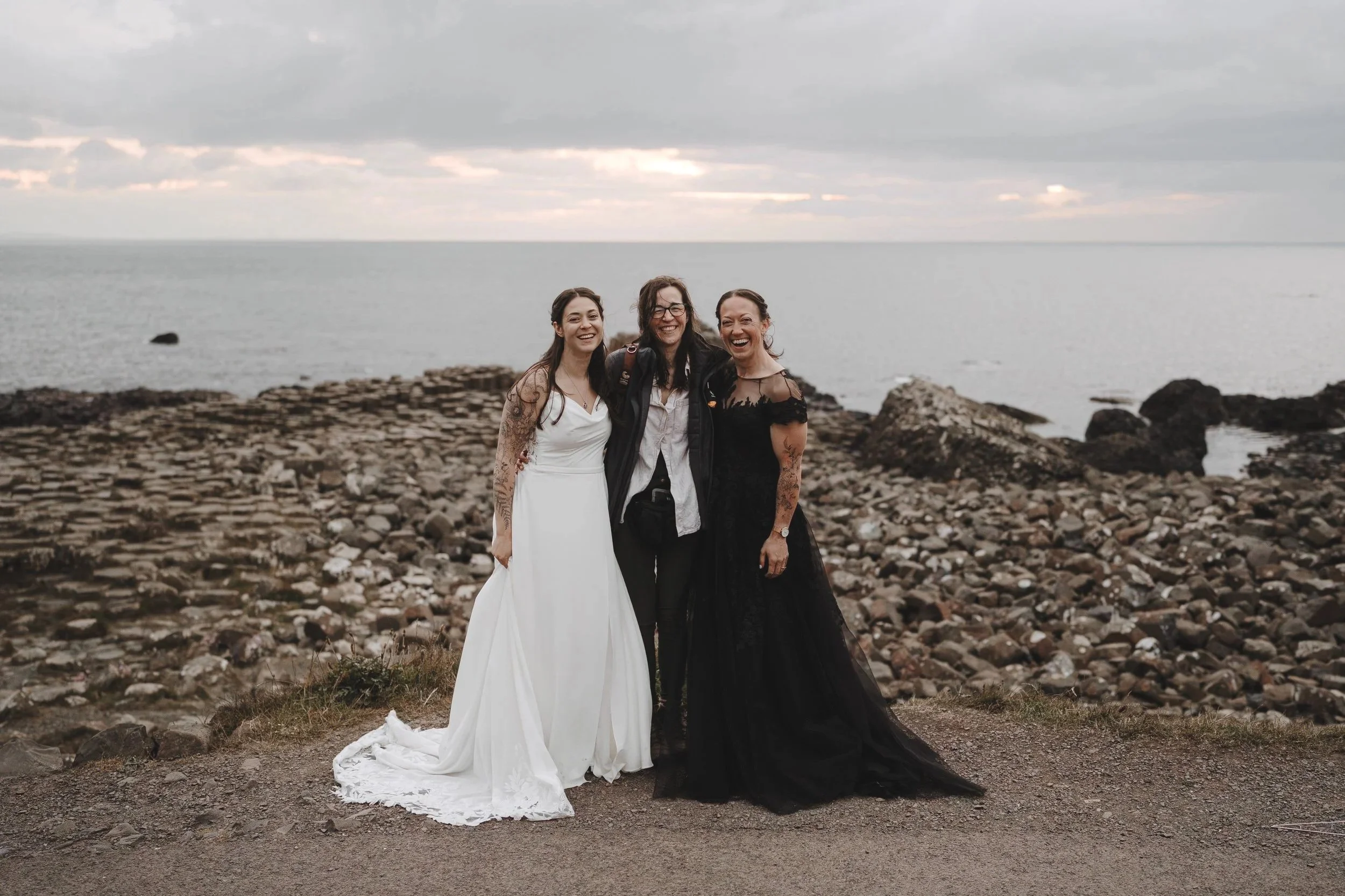 Three women standing on a rocky beach near the ocean, smiling. One is in a white dress, third in a black dress, middle in casual attire, with a cloudy sky overhead.