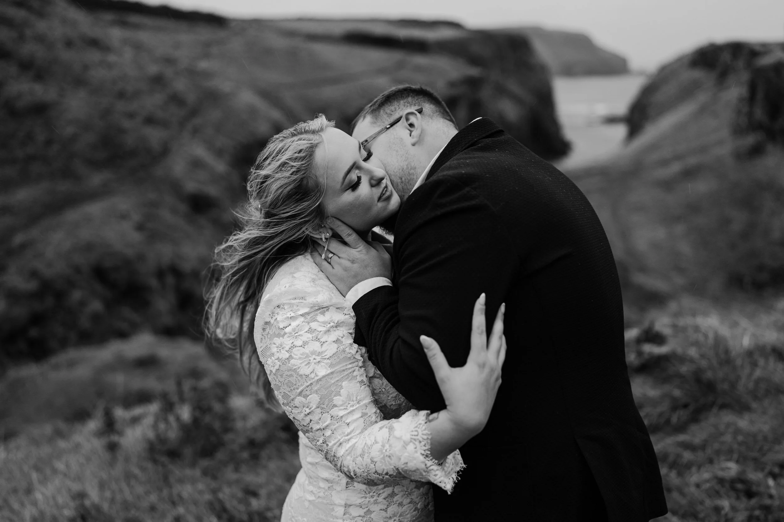 A black and white photo of a couple embracing outdoors, with rocky hills in the background. The woman is wearing a lace dress and the man is in a dark suit.