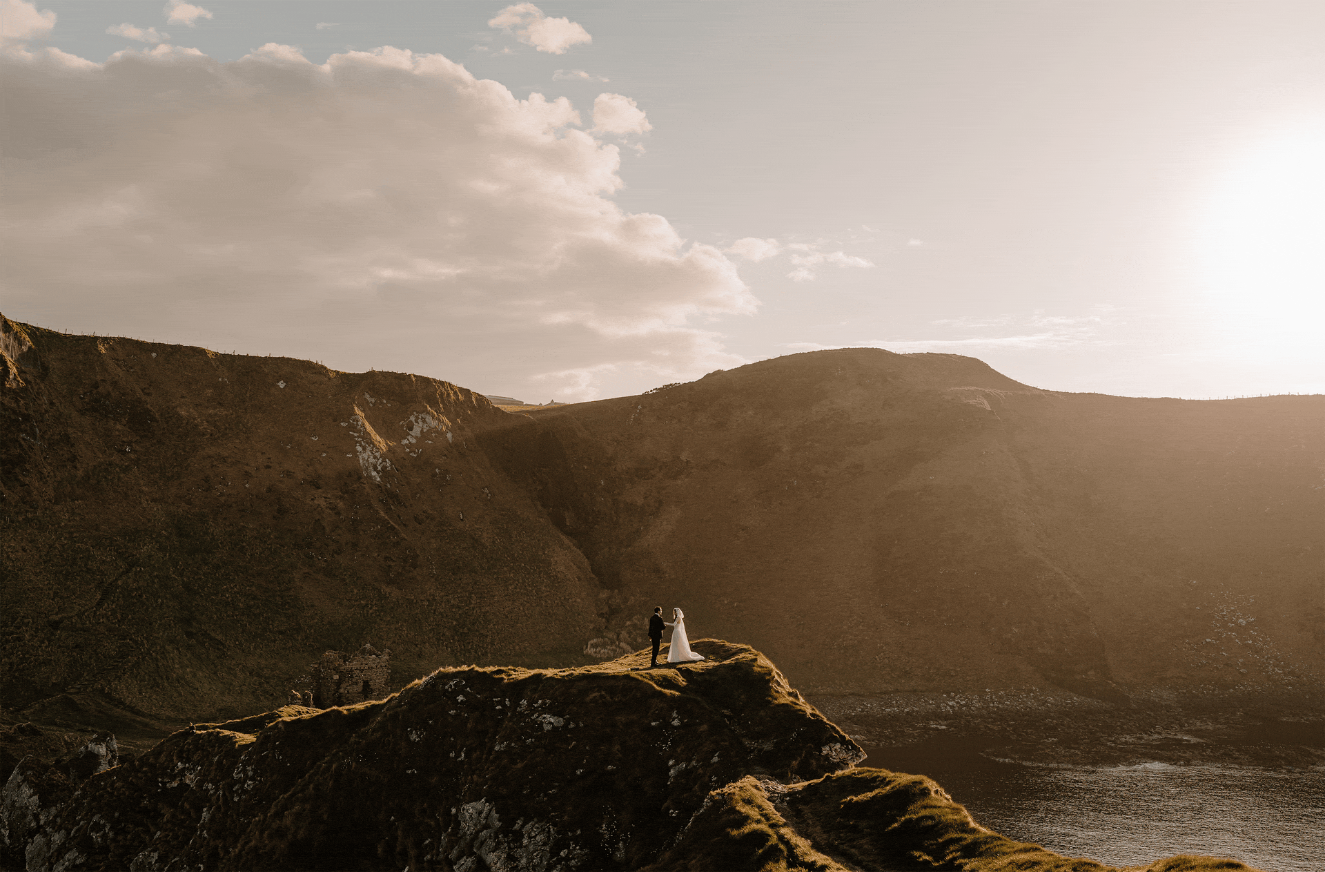 A bride and groom holding hands on a grassy hill at sunset with mountains and a cloudy sky in the background.