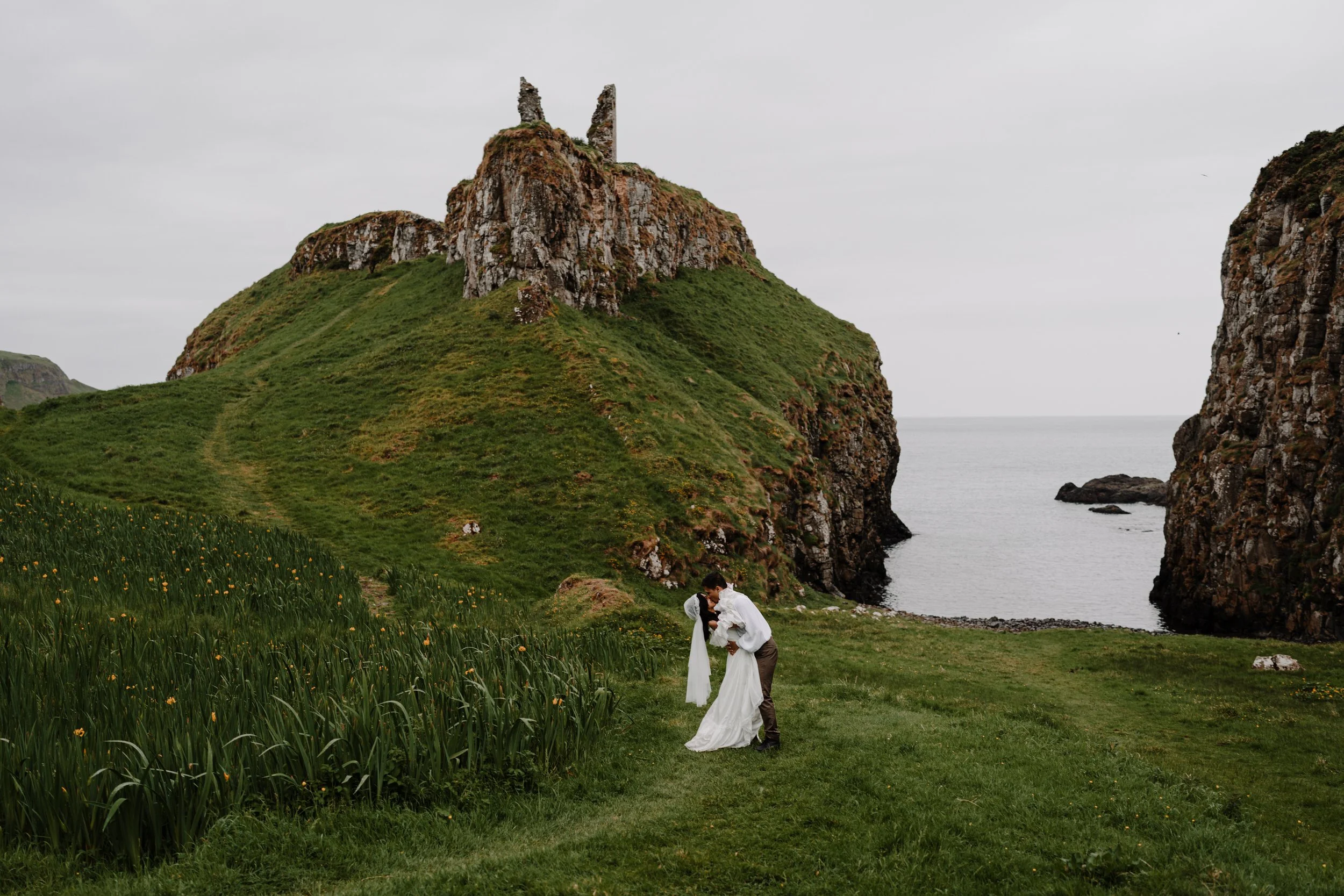 A romantic couple dressed in vintage attire, kissing on a lush green hillside with flowering grass, overlooking a rocky cliff and the ocean under a cloudy sky.