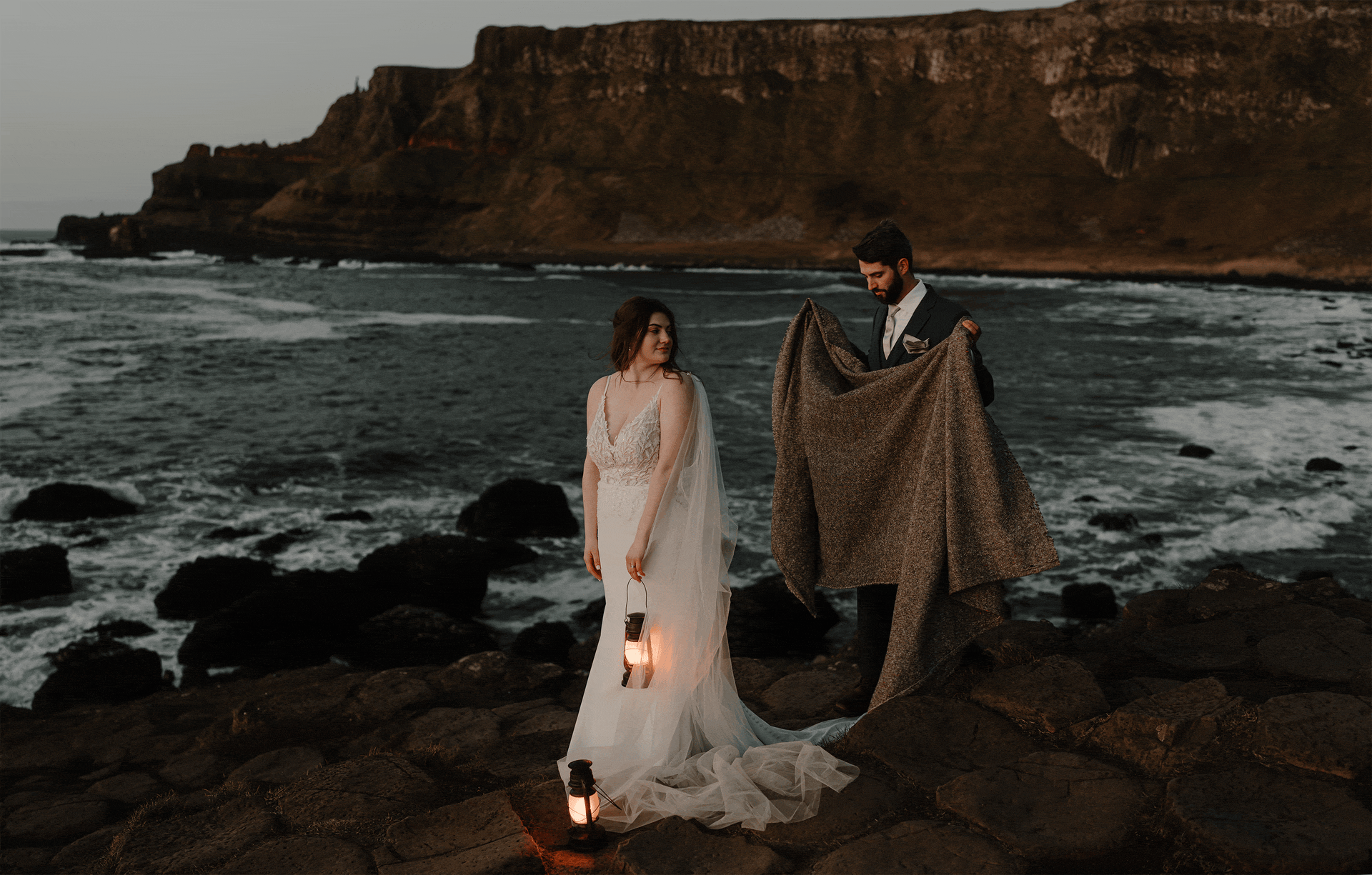 A couple dressed in wedding attire standing on rocks by the ocean at sunset, with cliffs in the background. The bride is holding a lantern and wearing a white gown, and the groom is holding a blanket.