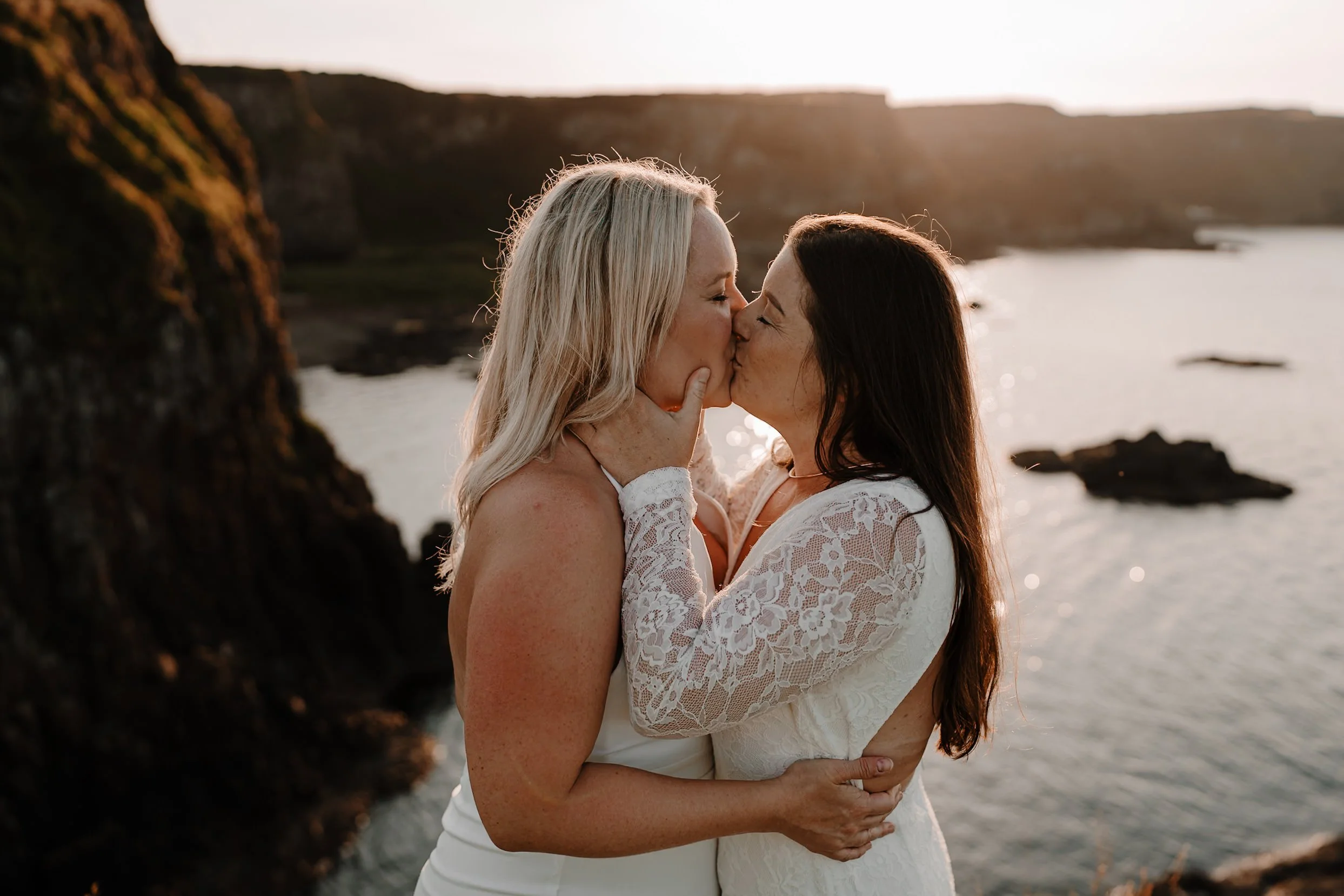 Two women in wedding dresses sharing a kiss at the beach during sunset, with a rocky shoreline and water in the background.