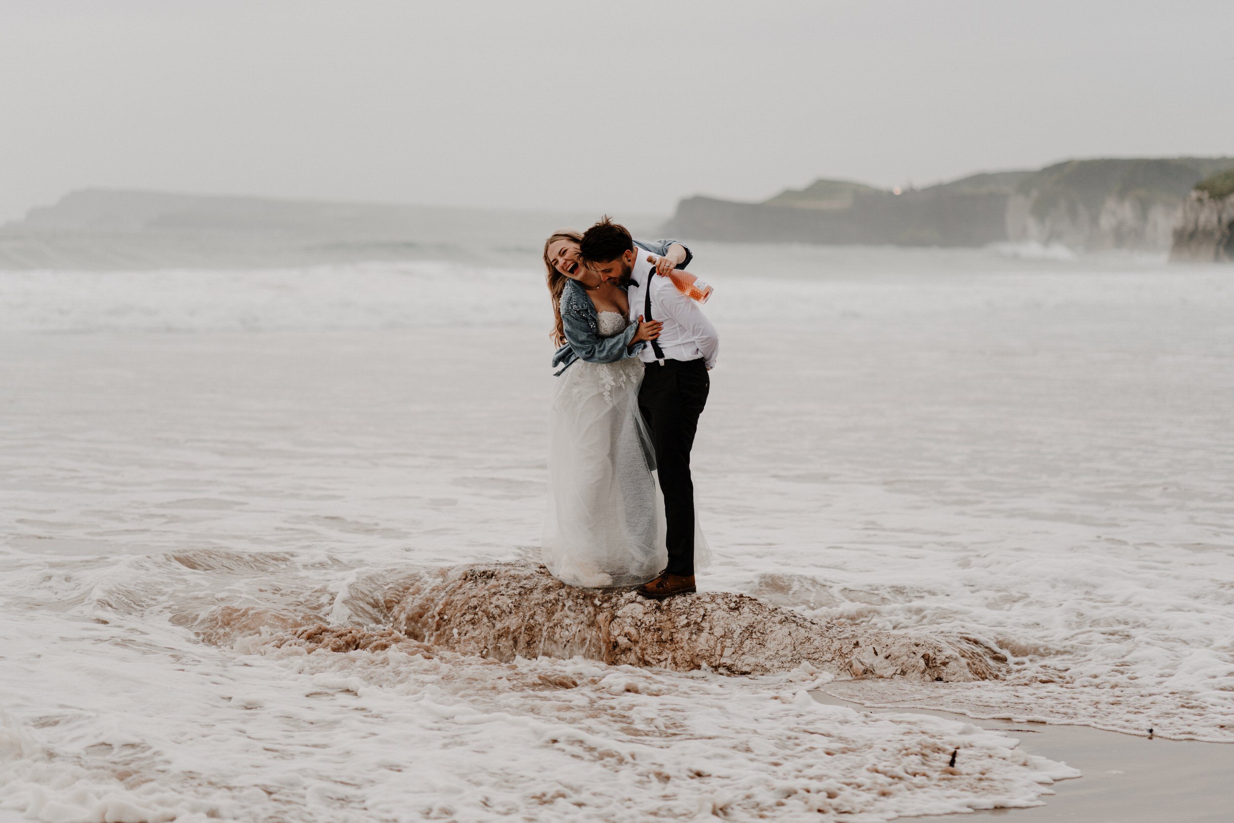 A bride and groom are standing on a rocky and sandy beach, embracing and laughing, with waves crashing around their feet and cliffs in the background on a cloudy day.