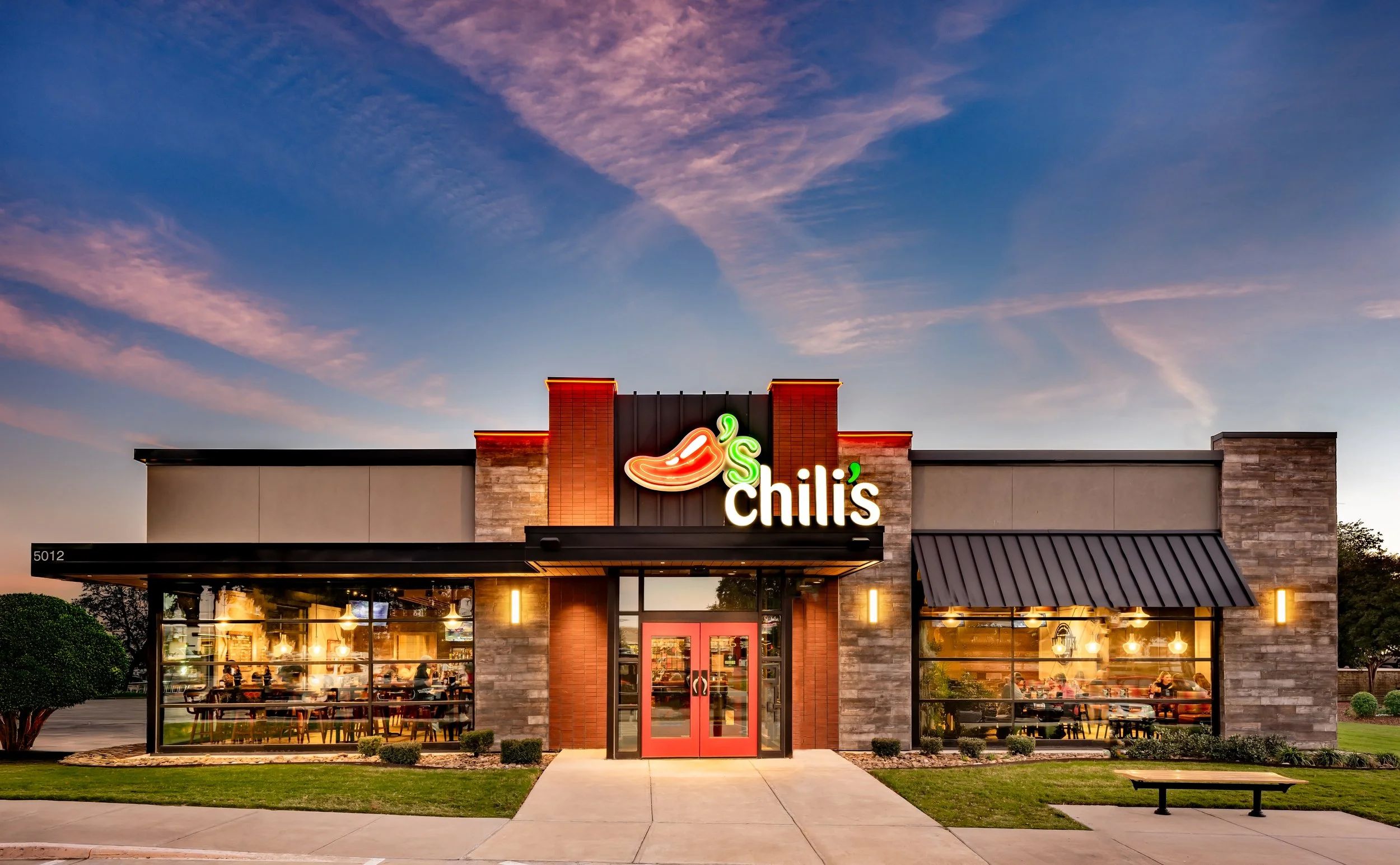 Exterior of Chili's restaurant during evening with illuminated sign and large windows showing dining area, under a partly cloudy sky.