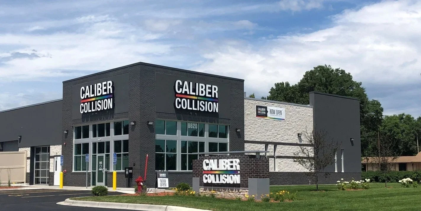 Exterior view of Caliber Collision auto repair shop with signage. Modern building with large windows, surrounded by landscaping and parking lot.