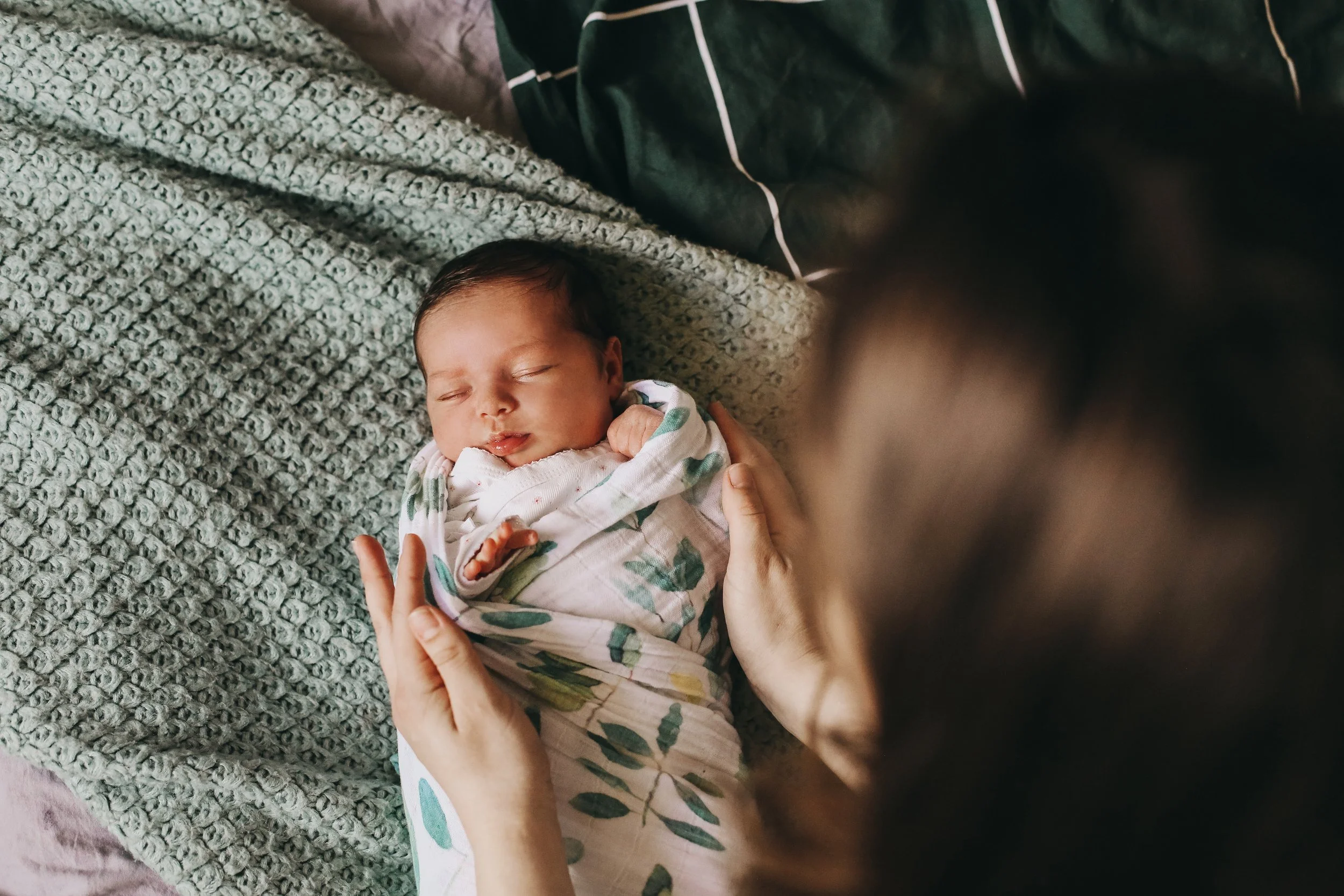 A newborn baby with dark hair sleeping on a textured gray blanket, gently held by an adult whose hand supports the baby's head and chest.