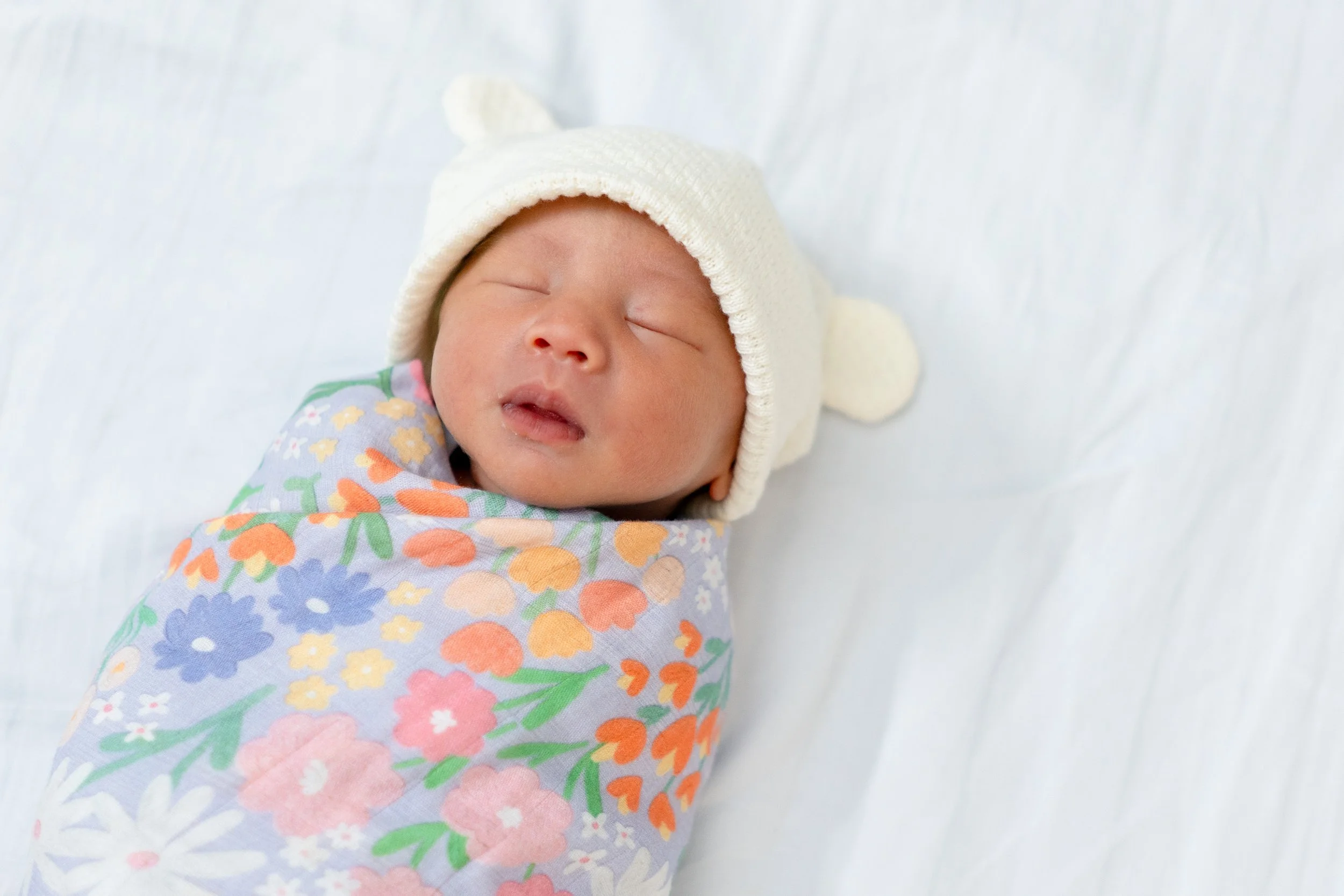 A sleeping baby wearing a white hat and a floral-patterned outfit lying on a white surface.