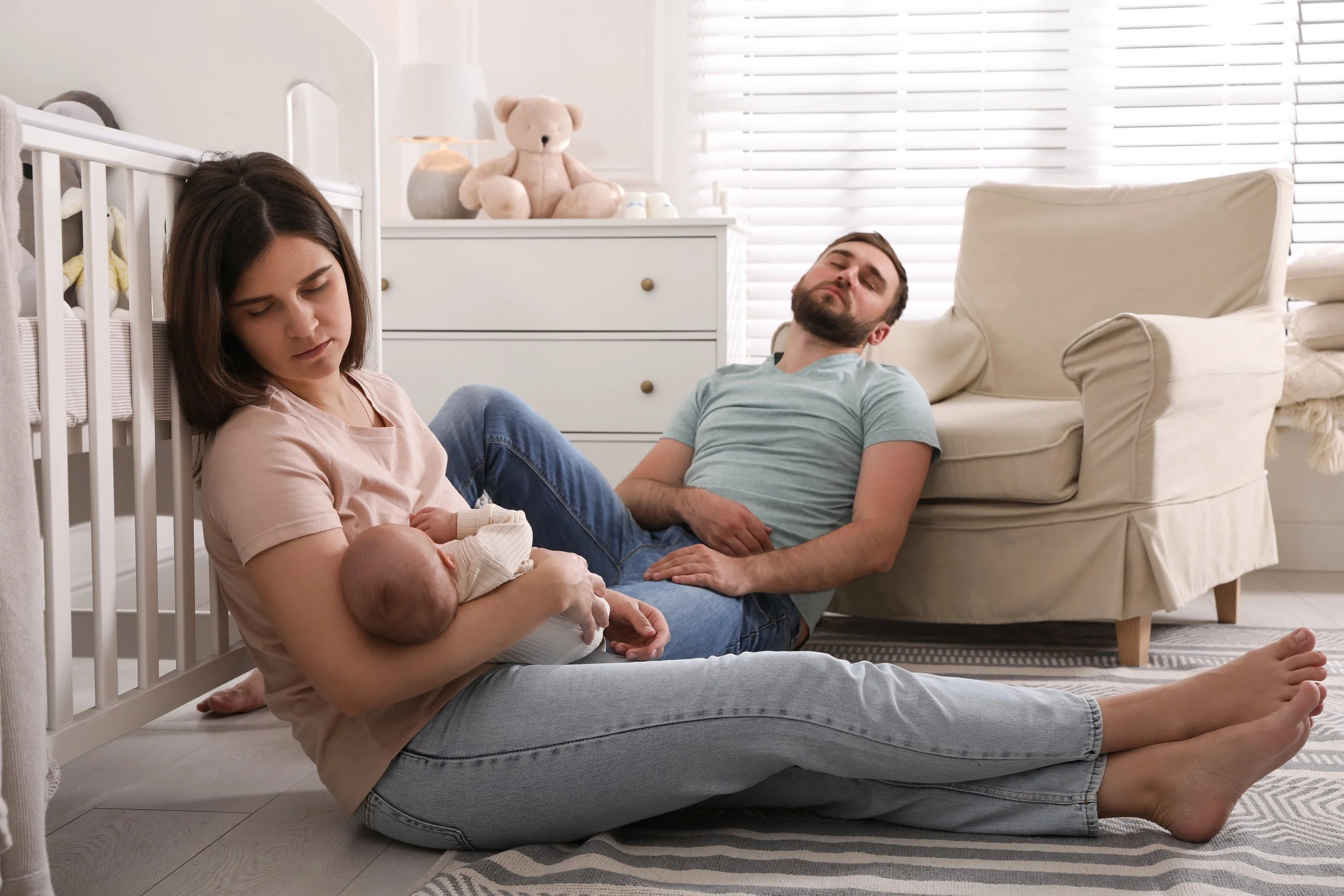 A young woman holding a newborn baby, sitting on the floor with her partner who is lying against a couch, in a bright nursery room.