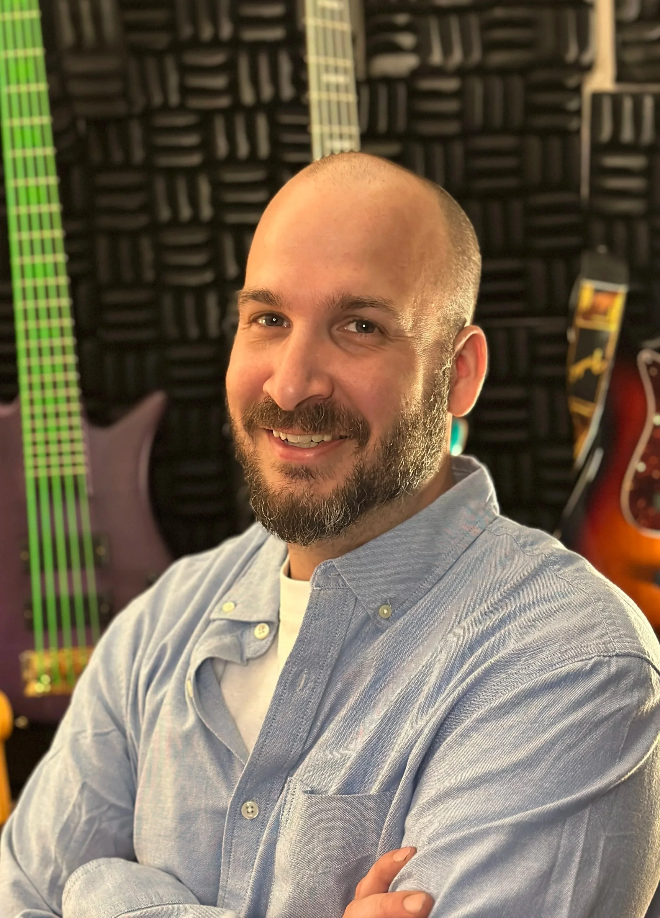 Kyle Kennedy smiles with arms folded wearing a blue bottun up and white tshirt. Behind him is a wall of colorful guitars