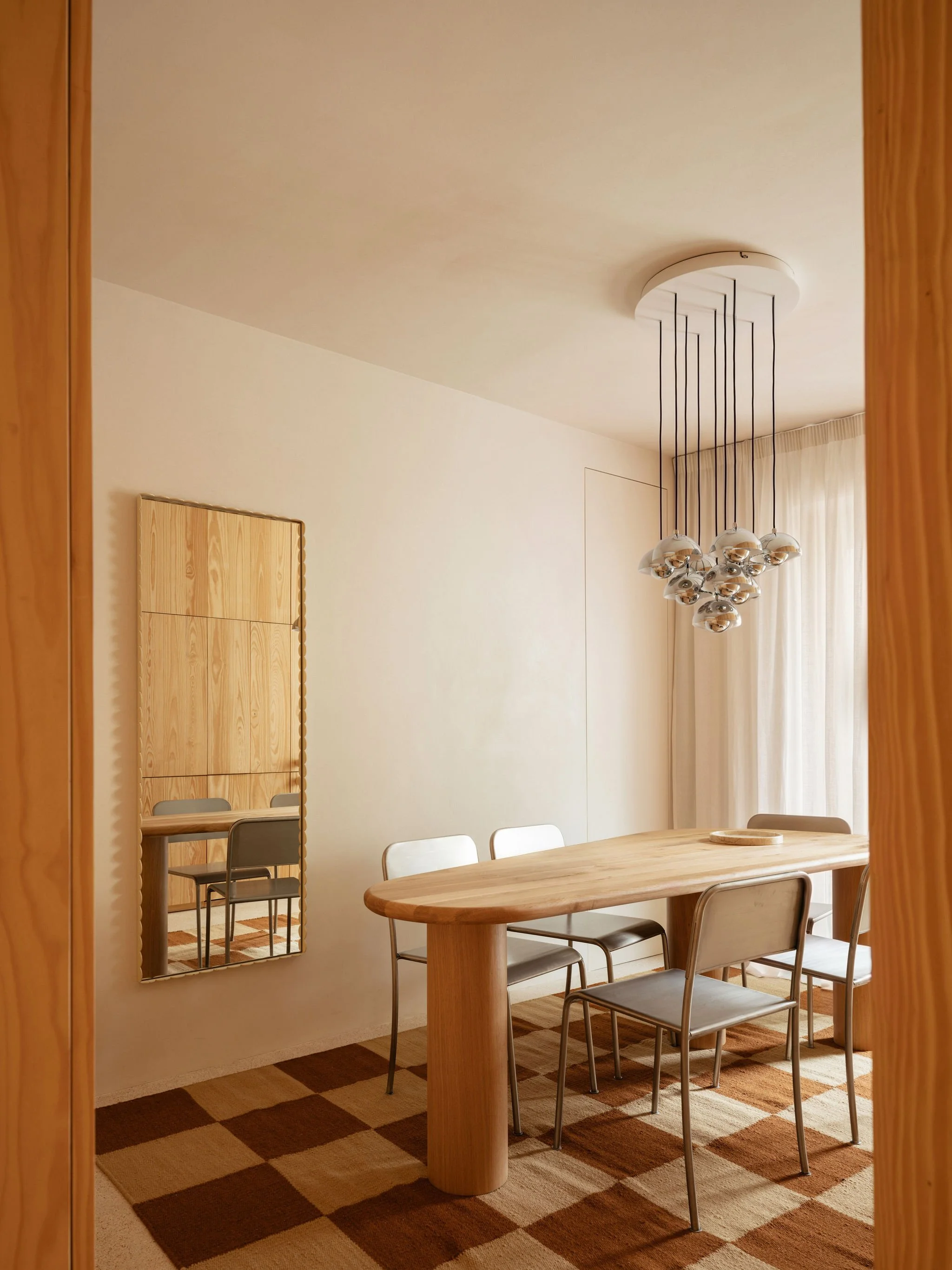 A dining room with a wooden table and six chairs, a floor mirror, a hanging chandelier, and a checkered carpet underneath.