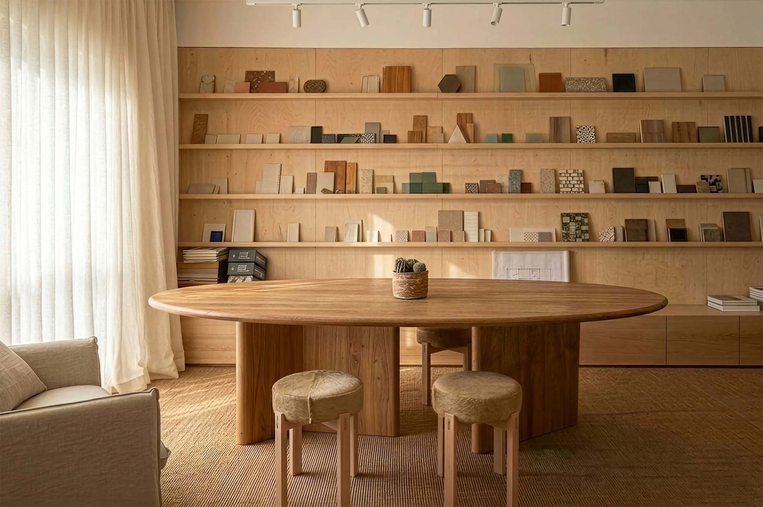 A room with a large wooden oval table, a small potted cactus on it, two beige stools, a beige armchair, and a wall of wooden shelving filled with small art pieces and books.