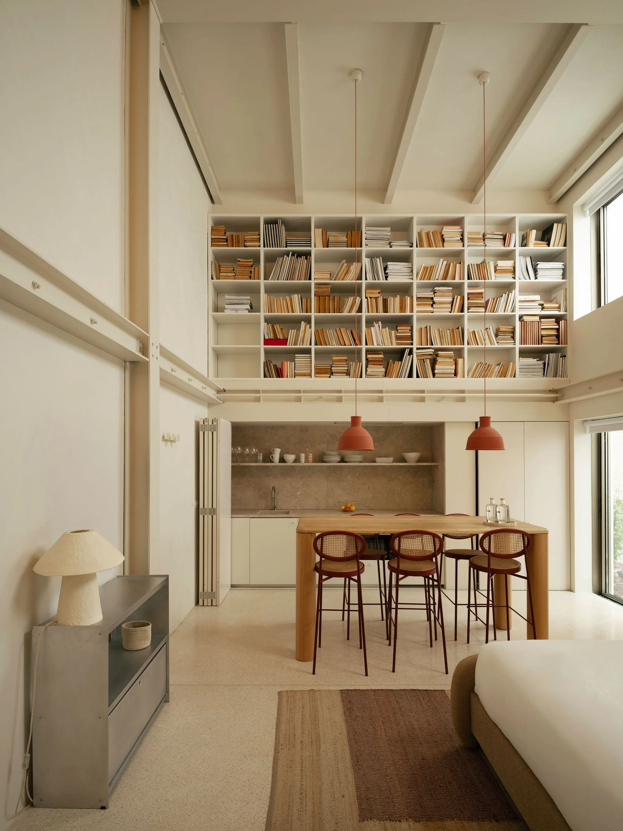 A modern, minimalist kitchen and dining area with a tall white bookshelf filled with books, a wooden dining table with four chairs, two pendant lights hanging above, a small counter, and large windows allowing natural light.