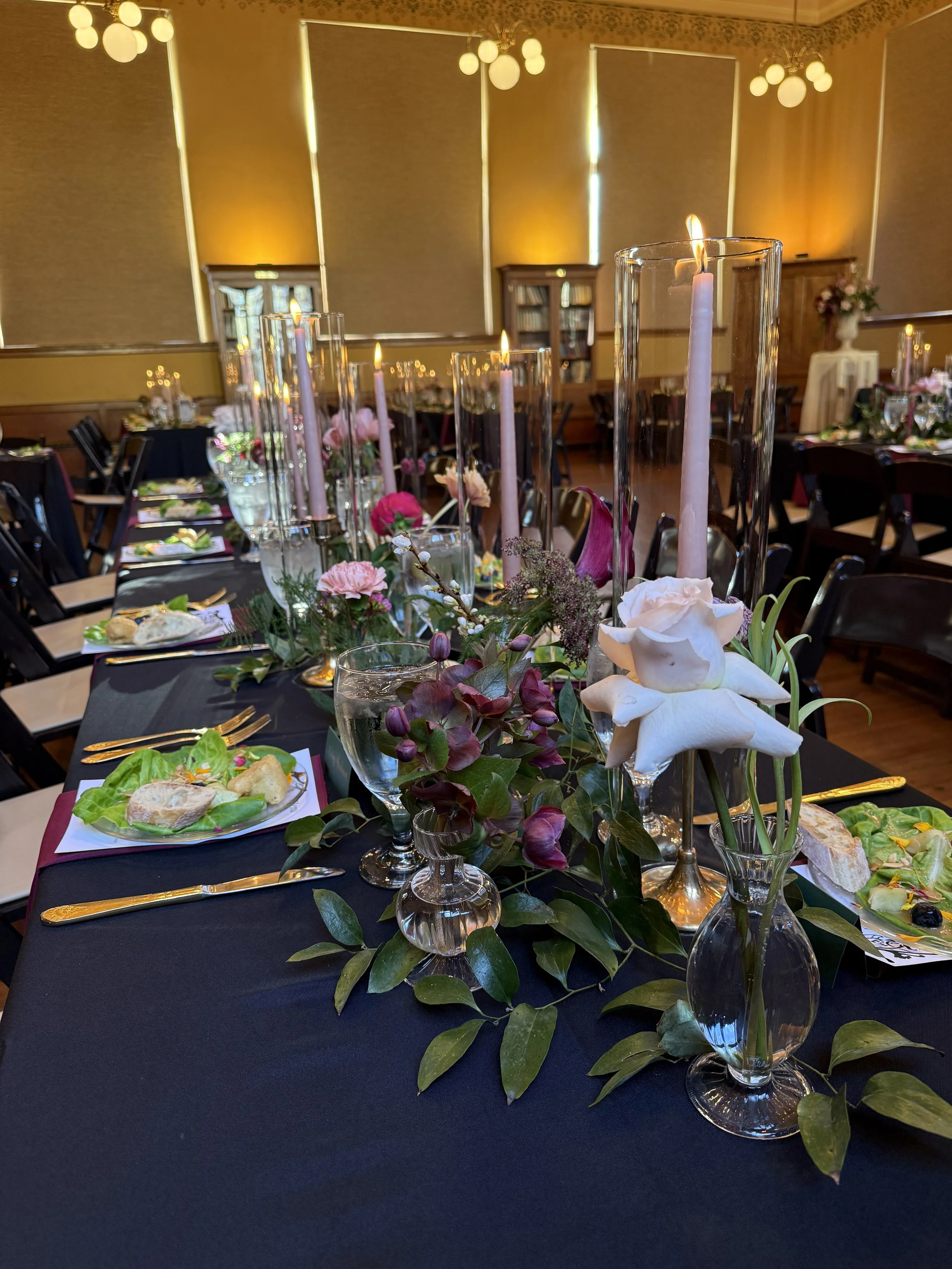 A dining table set for a formal event with black tablecloth, tall glass candle holders with white candles, floral arrangements with pink and white flowers, and plates of food with salad and bread. The room has warm lighting, beige walls, and windows 
