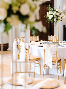 Elegant dining setup with white tablecloths, gold chairs, and a tall floral centerpiece of white roses and greenery.