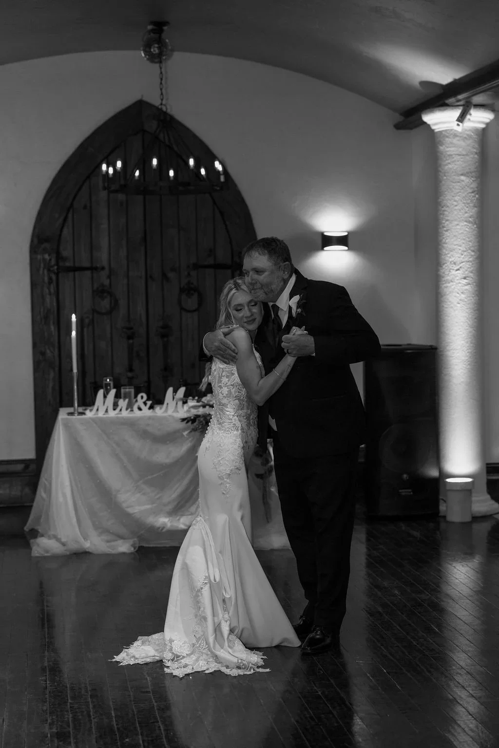 A bride and groom sharing their first dance at a wedding reception, with a decorated table and rustic backdrop in the background.