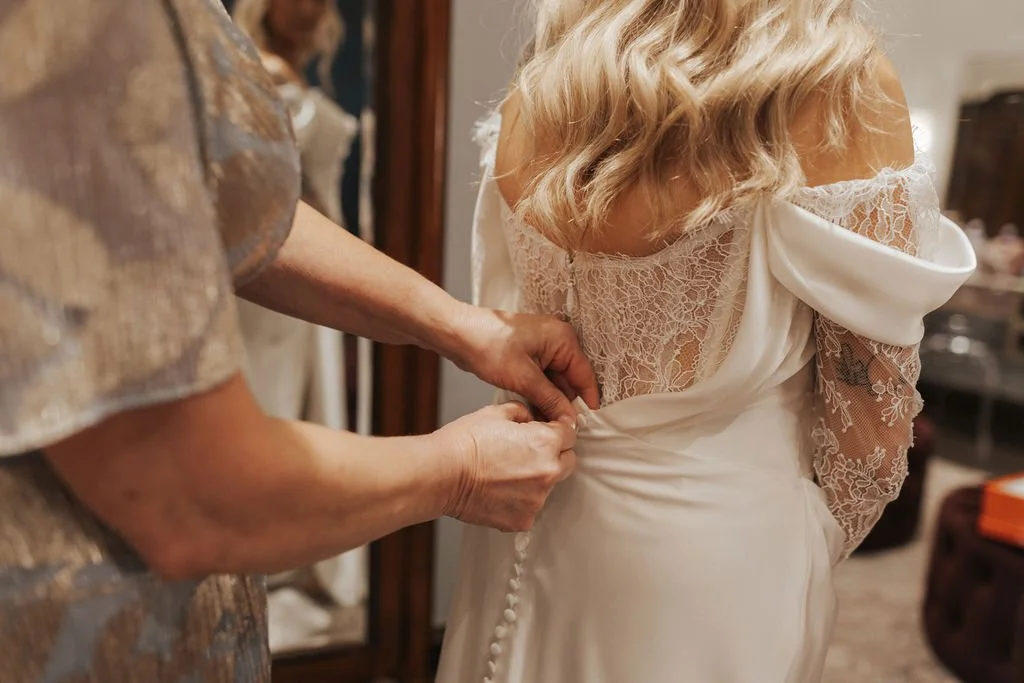 A timeless bridal moment—captured as the final buttons are fastened on her wedding gown. The perfect blend of elegance, emotion, and anticipation before walking down the aisle.