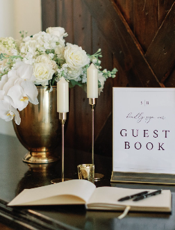 A guest book table with a large floral arrangement, a sign that says "Guest Book," an open guest book with a pen, and two white candles, all set on a dark wooden surface.