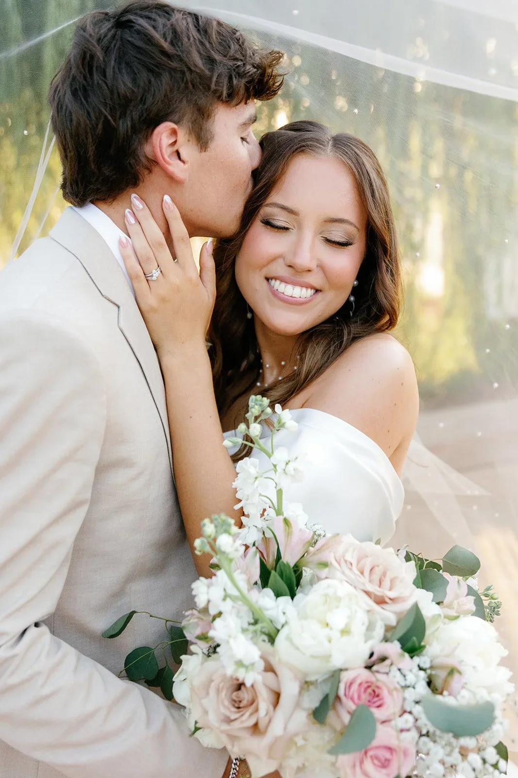 A bride and groom sharing a romantic moment on their wedding day, with the groom kissing the bride's forehead and the bride smiling with her eyes closed, holding a bouquet of flowers.