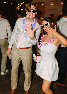 Two young adults at a party, wearing sunglasses and casual clothing, with string lights and other people in the background.