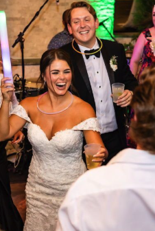 A bride and groom celebrating at their wedding reception, smiling and holding drinks, surrounded by guests.