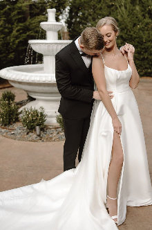 A bride and groom standing outdoors near a white fountain, with the groom leaning his head on the bride's shoulder as they smile. The bride is wearing a white wedding gown with a high slit, and the groom is in a black tuxedo.