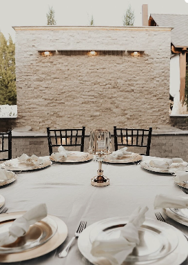 A set dining table outdoors with white tablecloths, plates, napkins, and glassware, in front of a beige brick wall with three small lights.
