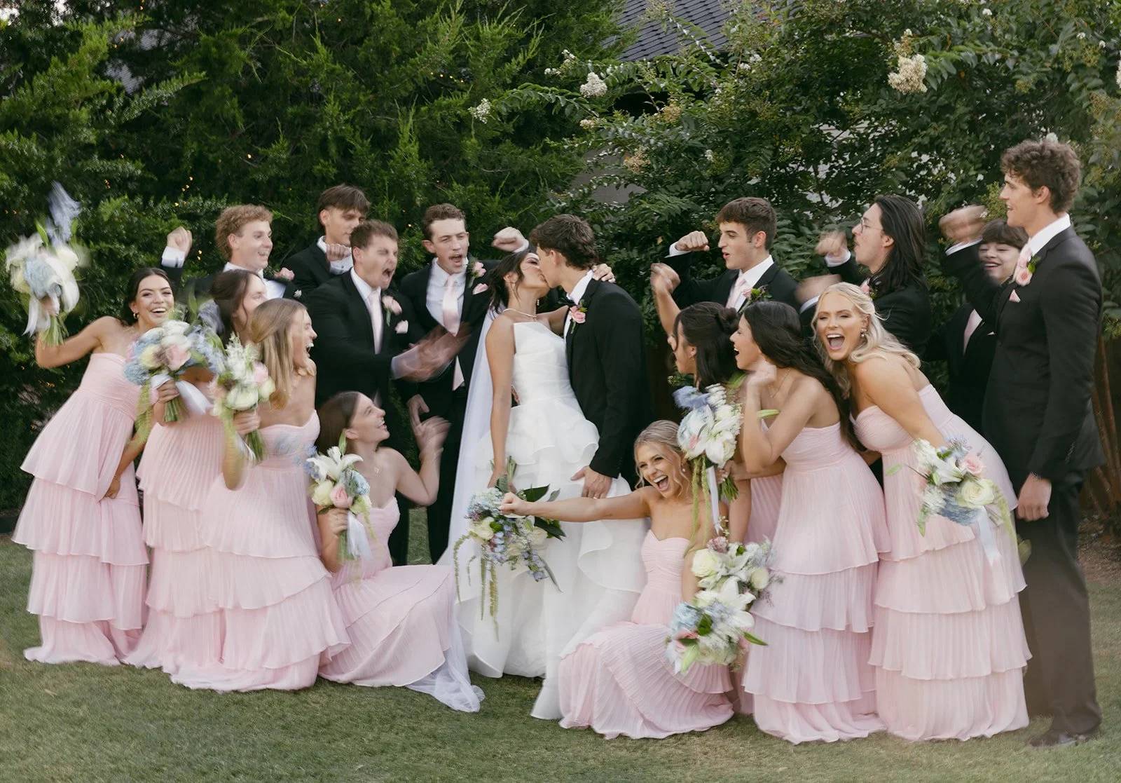 A group of people at a wedding, including a bride and groom kissing, surrounded by bridesmaids in pink dresses holding bouquets, and groomsmen in black suits, outdoors with green trees in the background.