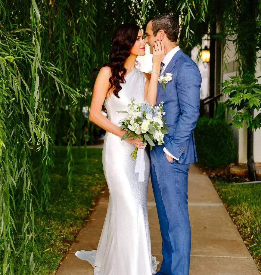 A bride and groom standing on a sidewalk under greenery, gazing into each other's eyes, with the bride holding a bouquet of white and blue flowers.