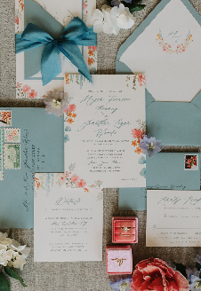 A collection of wedding invitations and envelopes decorated with flowers, a pink ring box, a red ribbon, and a blue bow.