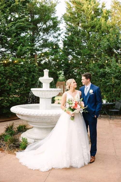 Bride and groom standing by a large outdoor fountain, surrounded by greenery and string lights, on their wedding day.