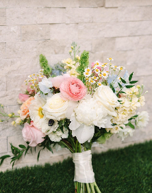A bouquet of various white, pink, and yellow flowers arranged with greenery, tied with a white ribbon, and placed against a brick wall.