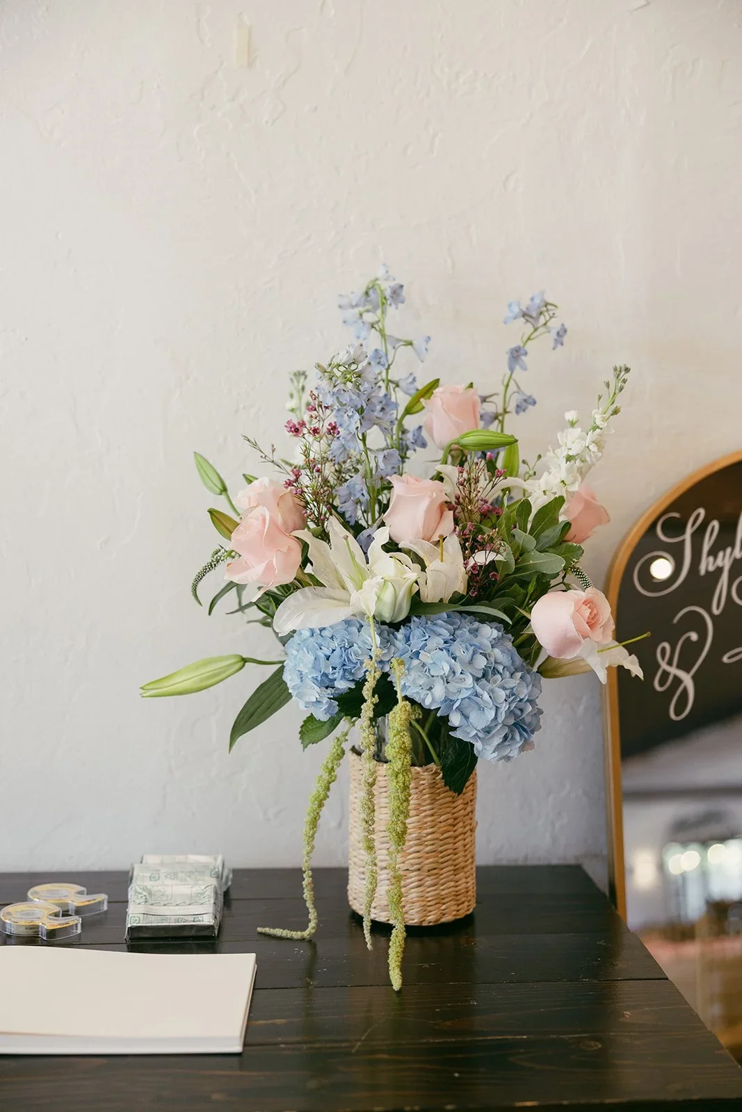 A bouquet of pink roses, white lilies, blue hydrangeas, and other flowers in a woven basket vase on a dark table.