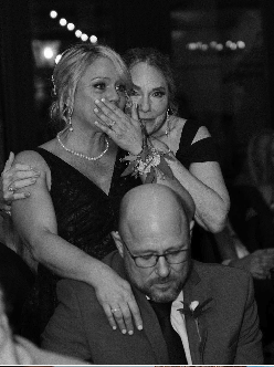 Three people at a social event, with two women standing behind a seated man. One woman appears surprised or emotional, covering her mouth with her hand, while the other woman looks at the camera. The man is looking down, wearing glasses.