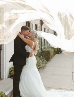 Bride and groom share a kiss under flowing white fabric on a city sidewalk.