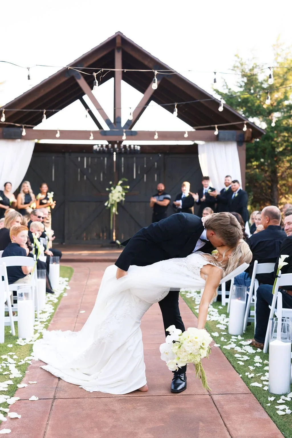 A bride and groom sharing a kiss during their wedding ceremony outdoors. The groom is leaning over the bride, who is bending backward, holding a bouquet of white flowers. Guests sit on either side of the aisle, and a barn-style structure with string lights hangs above.