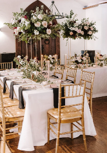 A decorated banquet table set for a special event with white tablecloths, gold chairs, and floral centerpieces, with hanging flower arrangements overhead.