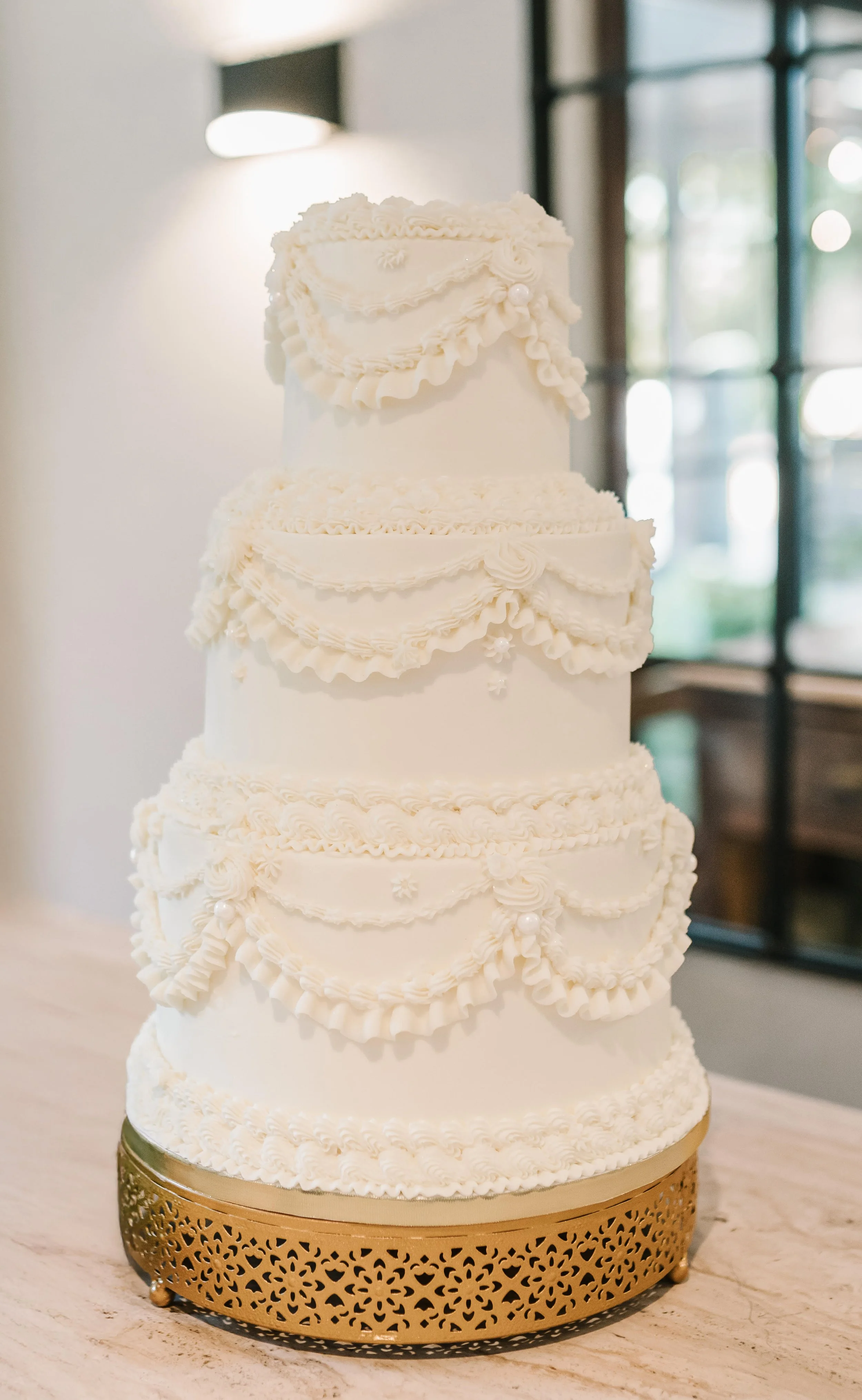 A multi-tiered white wedding cake decorated with intricate icing designs and ruffles, sitting on a gold decorative stand.