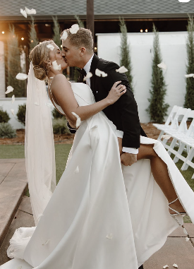 Bride and groom sharing a kiss outdoors during their wedding celebration, with falling flower petals and wedding chairs in the background.